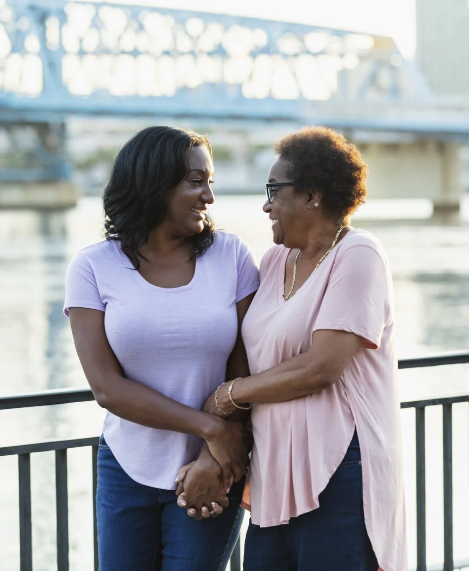 African American mother and adult daughter, arms linked and smiling on a bridge over water