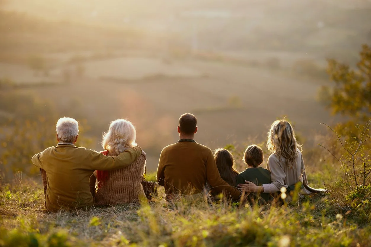 Multigenerational family sits on scenic hill, gazing at the sunset.