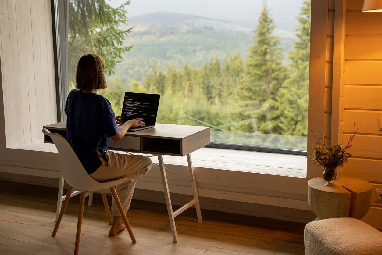 Person working on a laptop at a minimalist desk overlooking a green mountain landscape through a large window