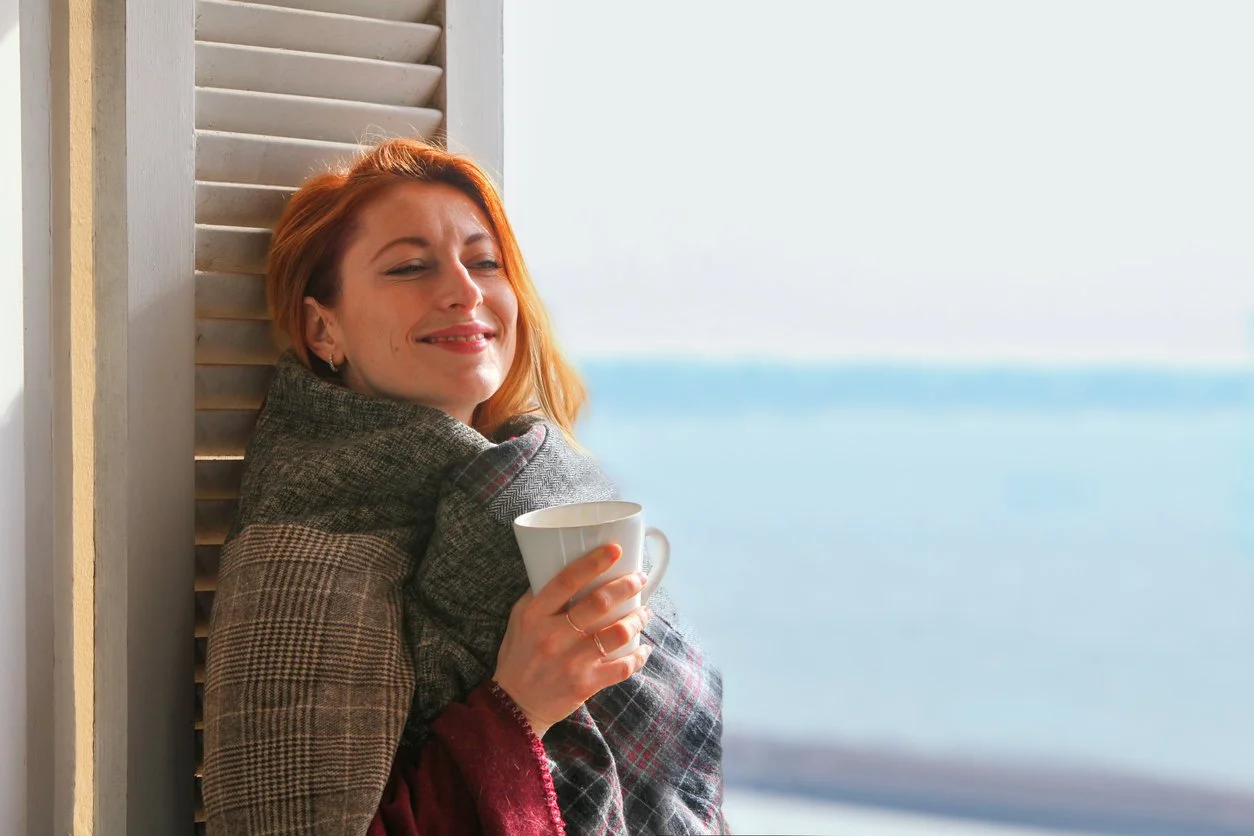 Red headed woman in 30s leaning in ocean view window, relaxed with tea.