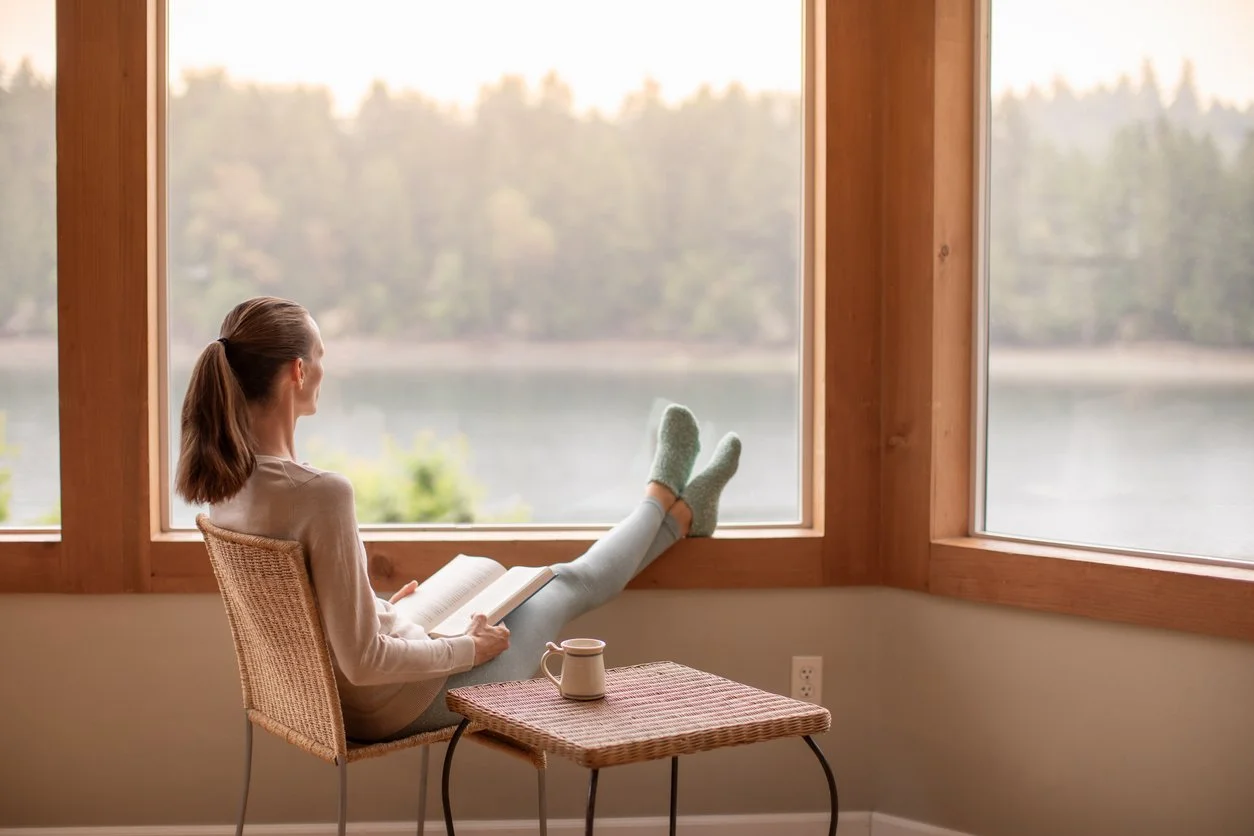 Woman relaxing with a book and coffee by a window overlooking a calm lake, feet up on a stool