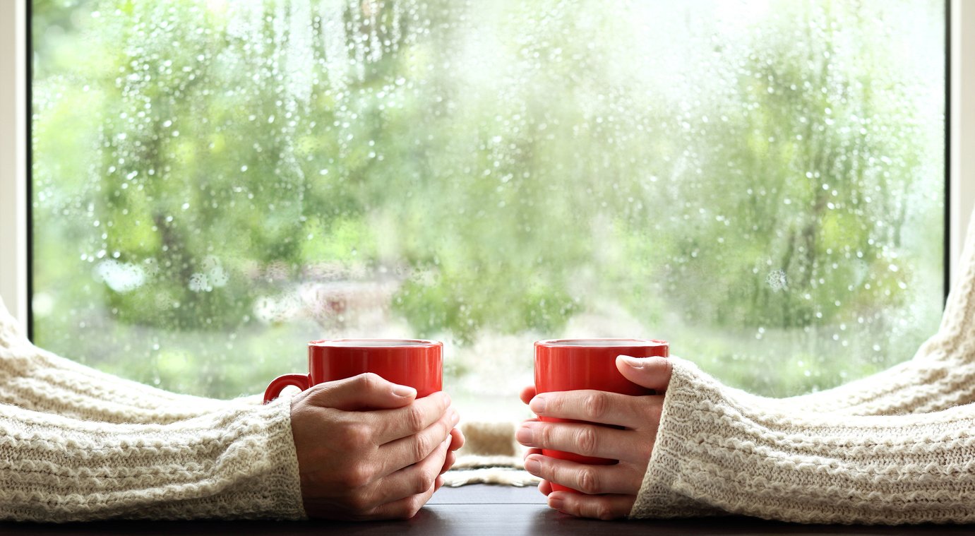 Two hands holding red mugs by a rainy window, cozy sweater sleeves — a warm, intimate conversation