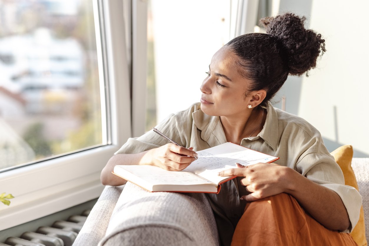 Young woman writing thoughtfully in a journal by a window, pen in hand