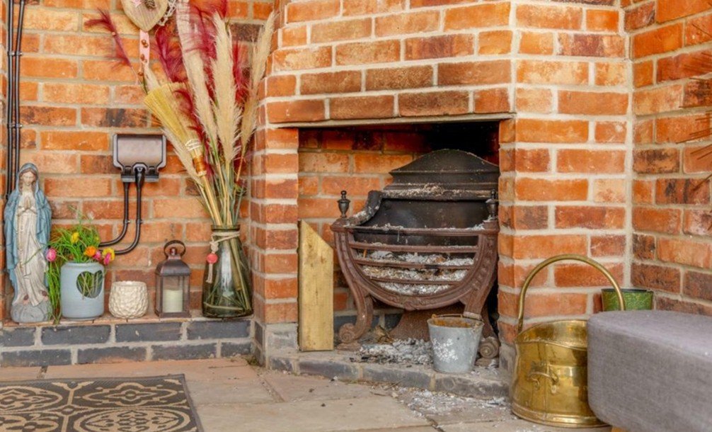 Brick fireplace with old, unused grate and ashes. Decor includes a religious statue, a vase with dried flowers, a lantern, and a gold-colored watering can placed on the floor.
