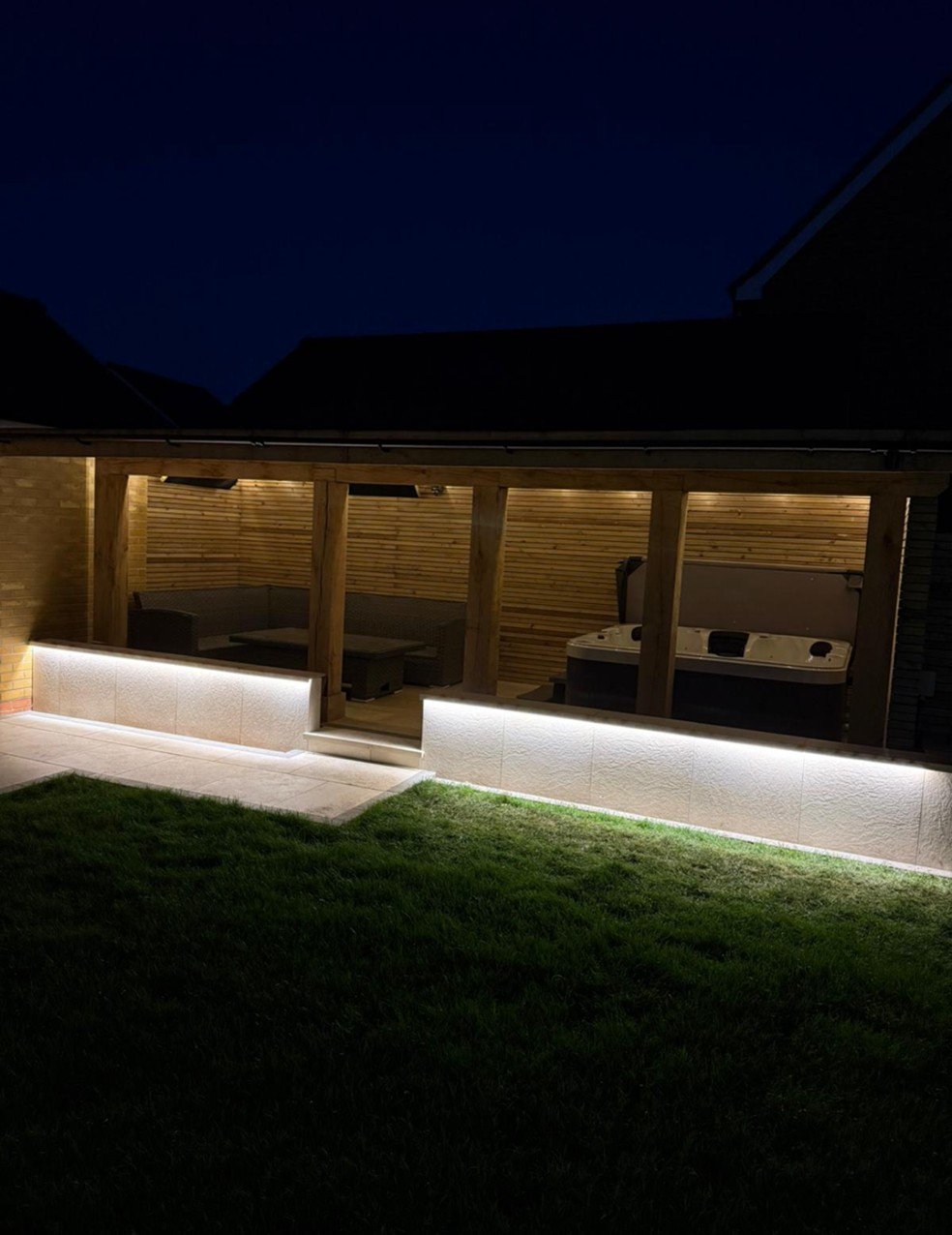Night view of a backyard patio with a hot tub, seating area, wooden pergola, and landscape lighting.