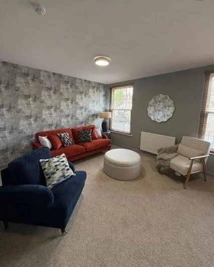 Living room with gray carpet, gray wall with textured wallpaper, red sofa, navy blue armchair, white ottoman, beige armchair, window showing trees outside, wall mirror, lamp on side table, ceiling light