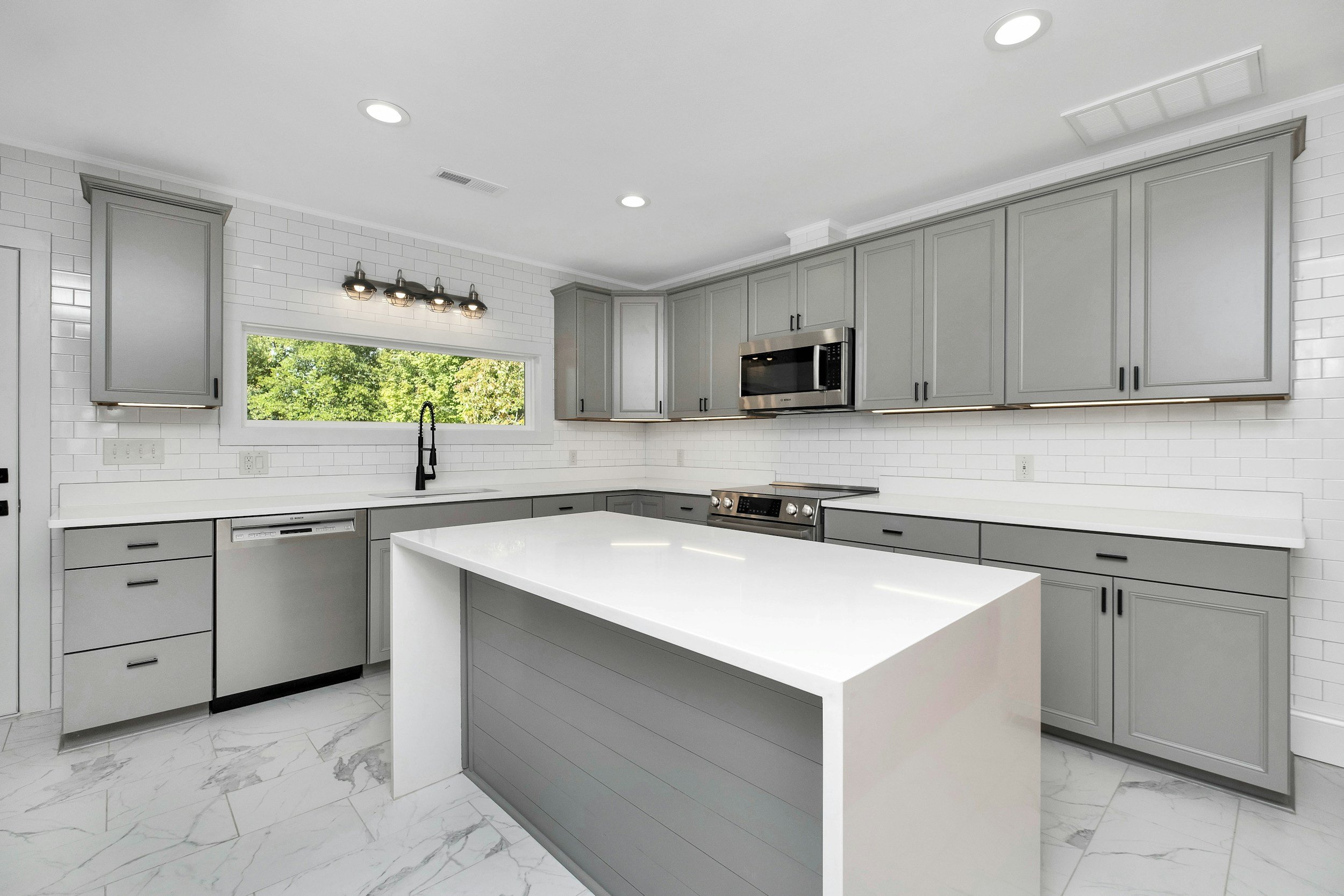 Modern kitchen with gray cabinets, white countertops, stainless steel appliances, a large island, white subway tile backsplash, and a window with view of green trees.