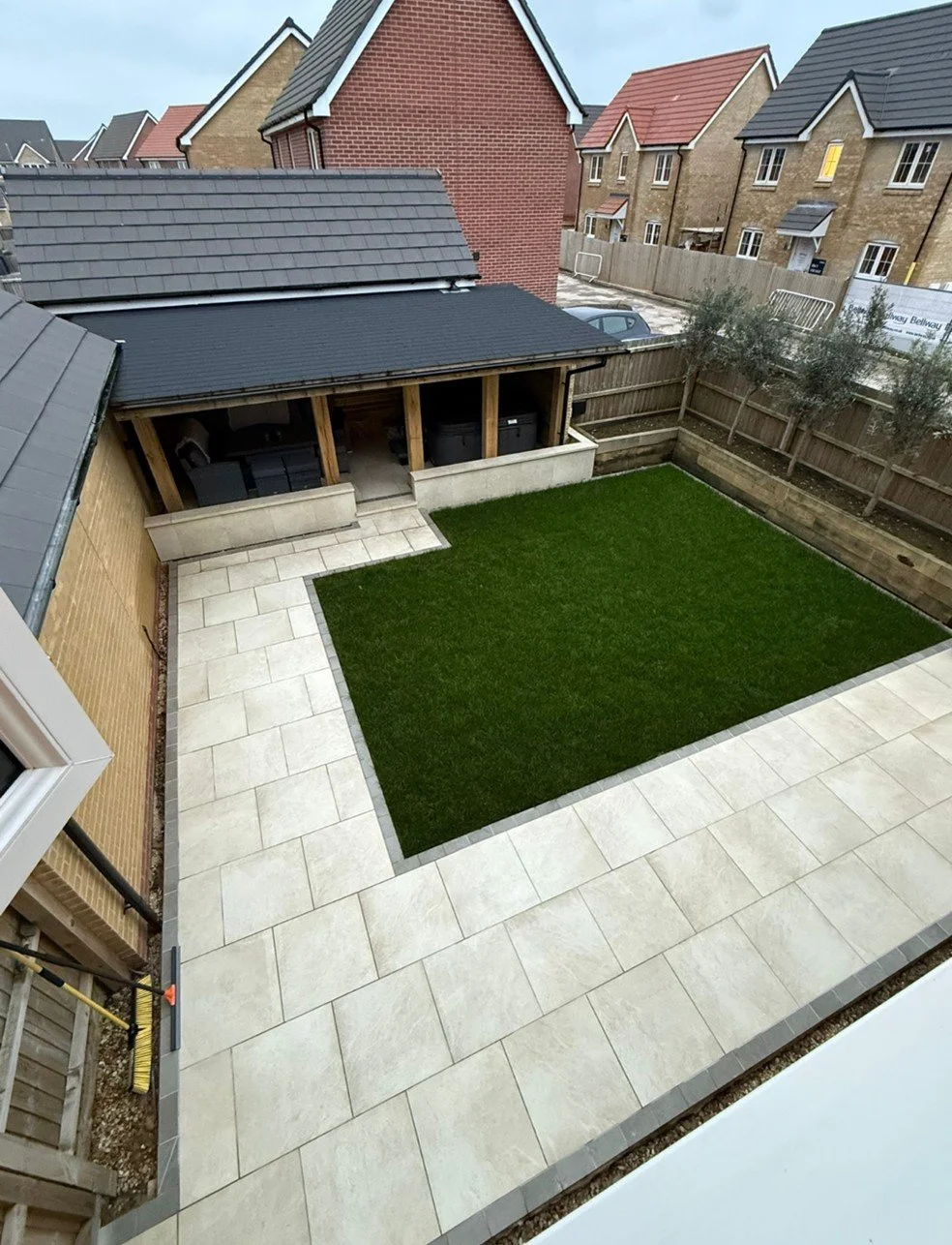 View of a backyard with a small grassy area, paved stone patio, and a covered outdoor seating area with chairs and storage containers, surrounded by a wooden fence.
