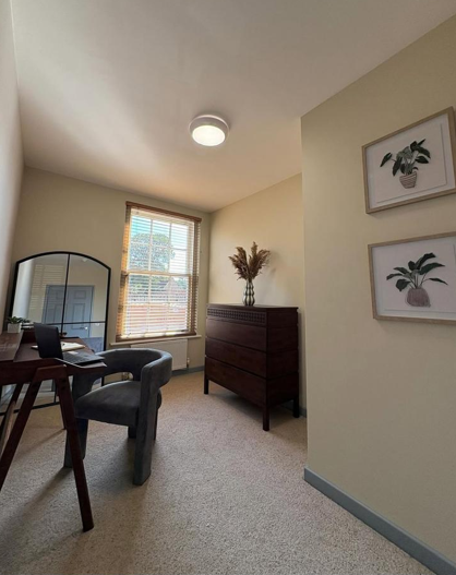 A cozy home office with a window, a wooden desk, an upholstered chair, framed botanical prints on the wall, and a dresser with decorative dried flowers.