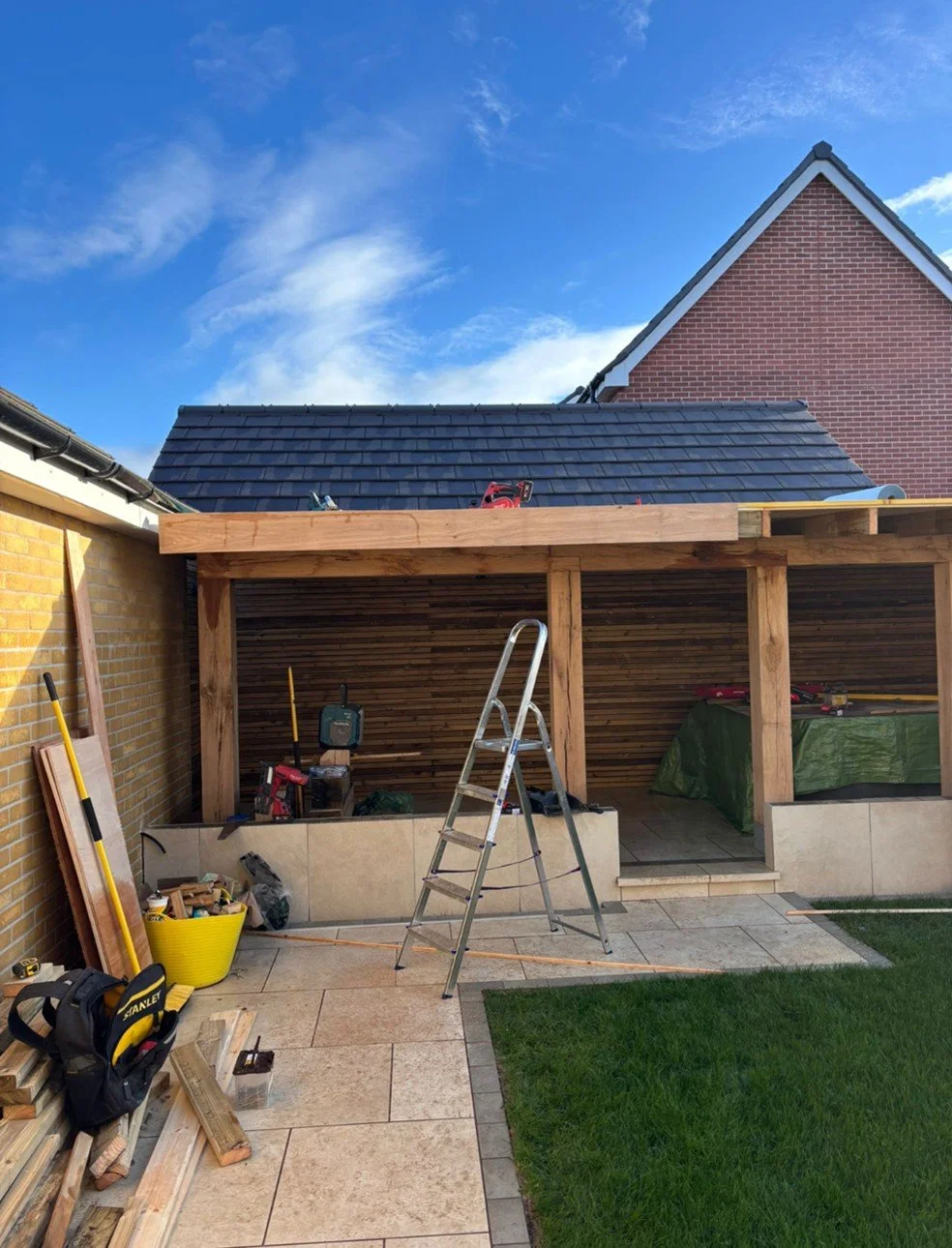 A backyard construction site with a wooden structure being built, tools and materials scattered around, and a ladder in front. The sky is blue with some clouds, and neighboring houses with brick walls and tiled roofs are visible.