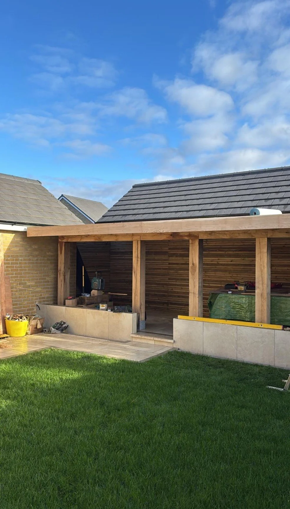 Construction of an outdoor patio or extension with wooden beams and walls on a backyard with lush green grass under a partially cloudy sky.