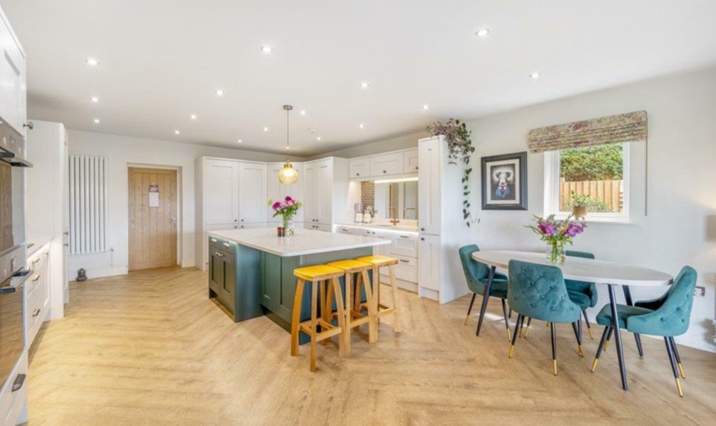 Modern open-concept kitchen and dining area with white cabinets, a green kitchen island, wooden stools, a round dining table with teal chairs, and flowers on the table and island.