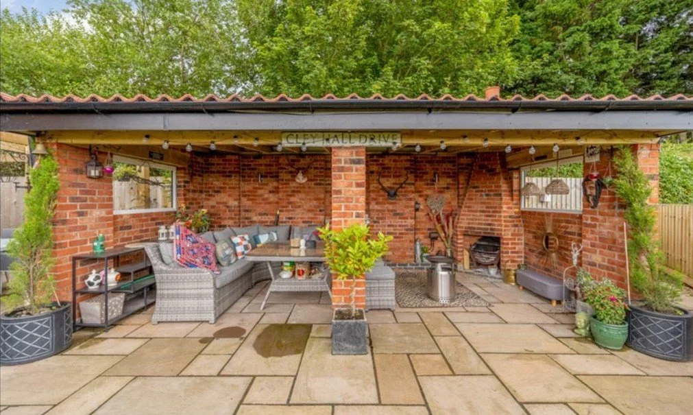 Outdoor patio area with brick walls, a seating area with cushions, potted plants, and decorations, under a roof with string lights.