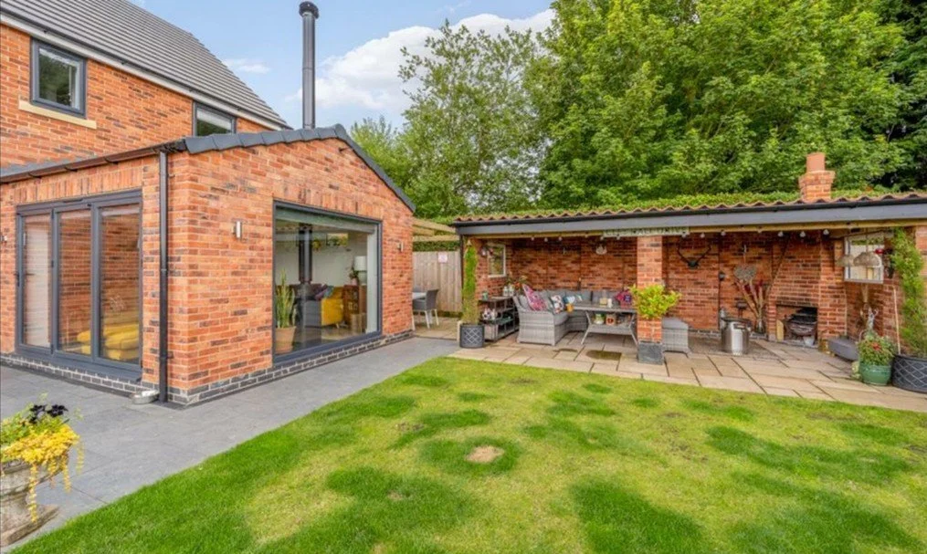 Backyard with brick house extension, patio area with outdoor seating, covered brick barbecue area, green lawn, and trees in the background.