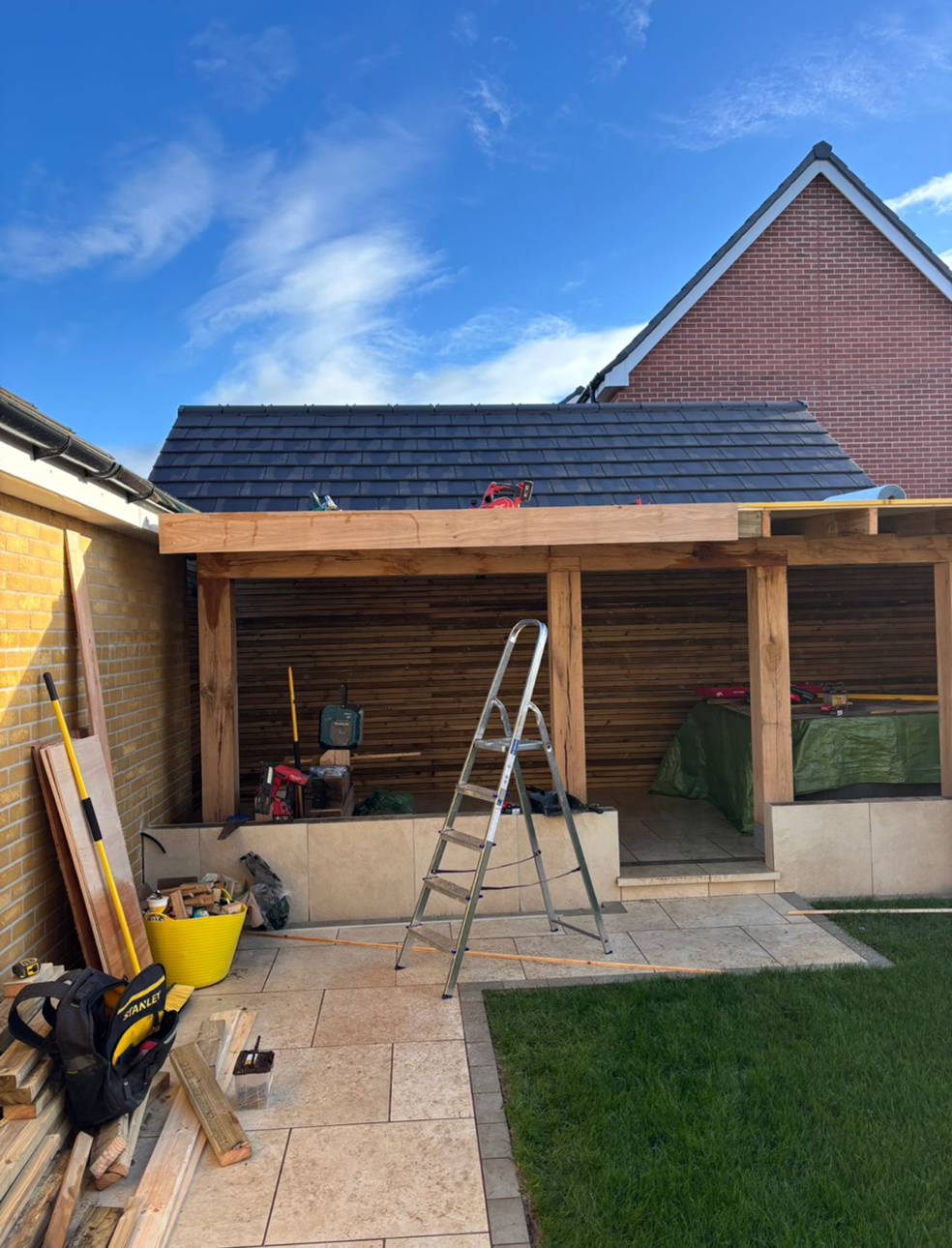 Construction of an outdoor wooden structure with tools and materials, with a brick house and blue sky in the background.