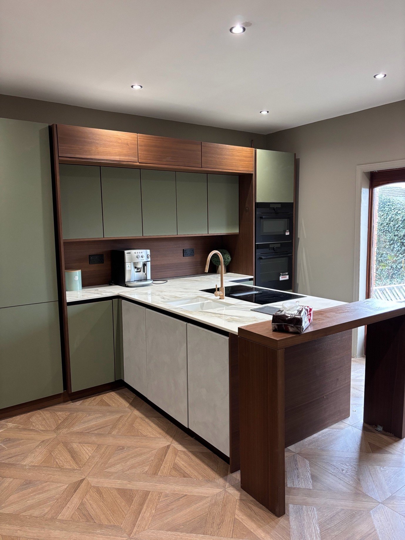 Modern kitchen with green cabinets, a marble countertop, black appliances, and wooden accents. Contains a coffee machine, sink, and a window with view outside.