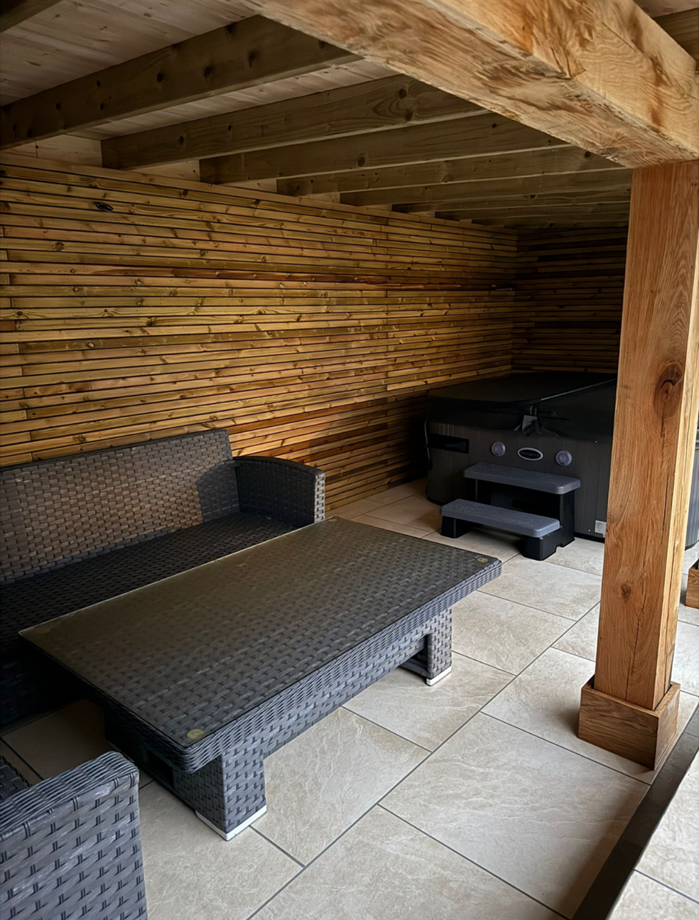 Outdoor patio with wicker furniture, a rectangular table, and a hot tub against a wooden slat wall and ceiling.