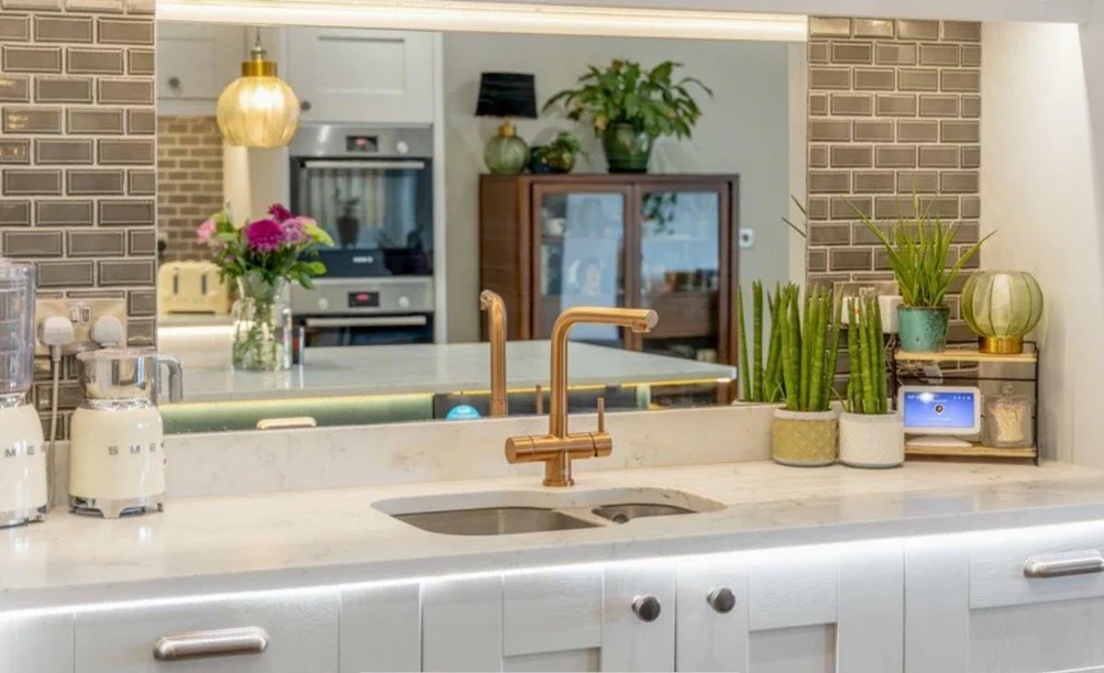 Kitchen with white countertop, gold faucet, green plants, and decorative items, with a view into the living area and brick walls.