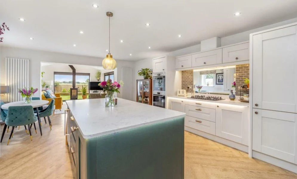 Open-concept kitchen and dining area with white cabinets, a large kitchen island with a pendant light, a dining table with teal chairs, and a living space with a view of a backyard.