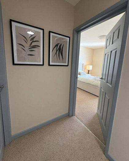 View of a hallway leading into a bedroom with a bed, lamp, and nightstand. Two framed botanical prints hang on the hallway wall.