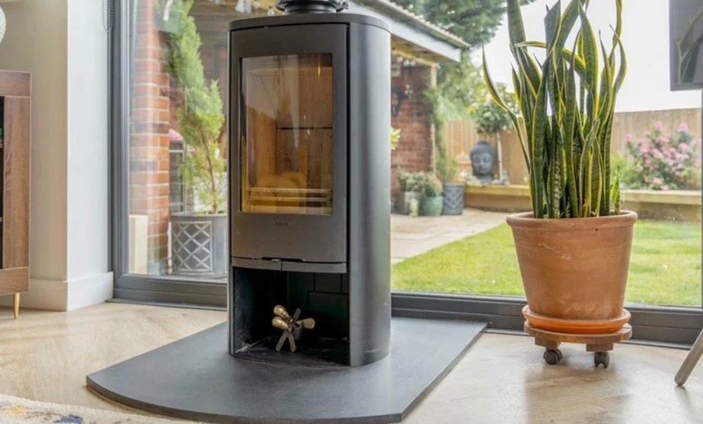 A modern black freestanding wood stove on a curved hearth inside a home, with large glass windows showing an outdoor garden with potted plants, a Buddha statue, and flowering bushes.