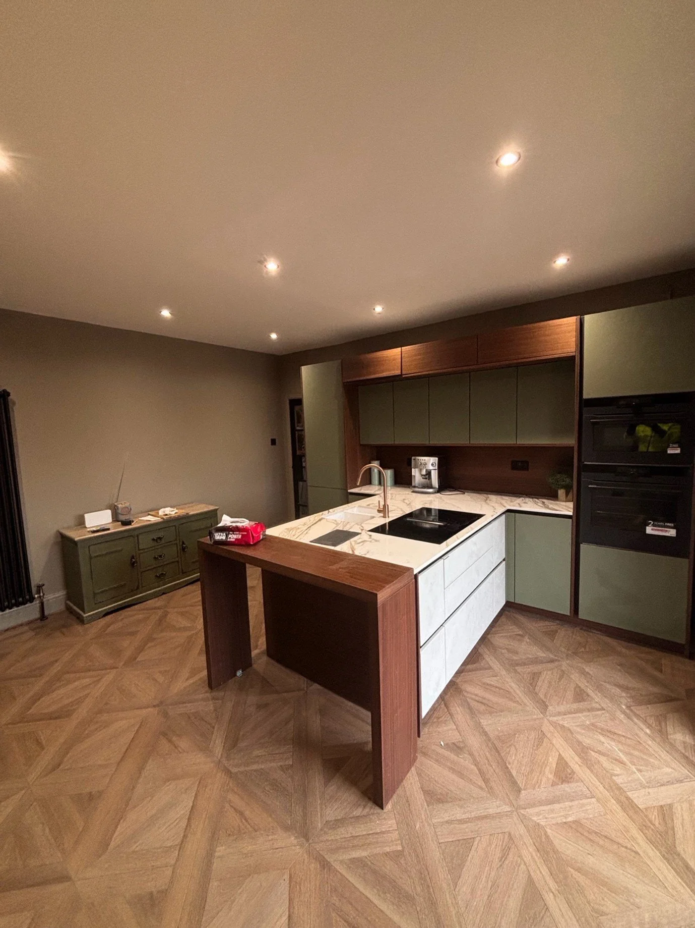 Modern kitchen with green cabinets, a marble island, wooden accents, and a herringbone wood floor.
