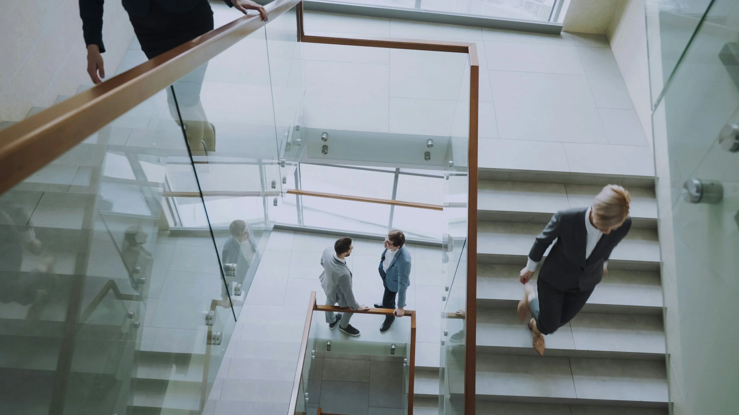 A view from the top looking down at a modern office building staircase with glass balustrades. There are three people visible: two are standing and talking at the bottom of the stairs, and one person is walking up the stairs on the right.