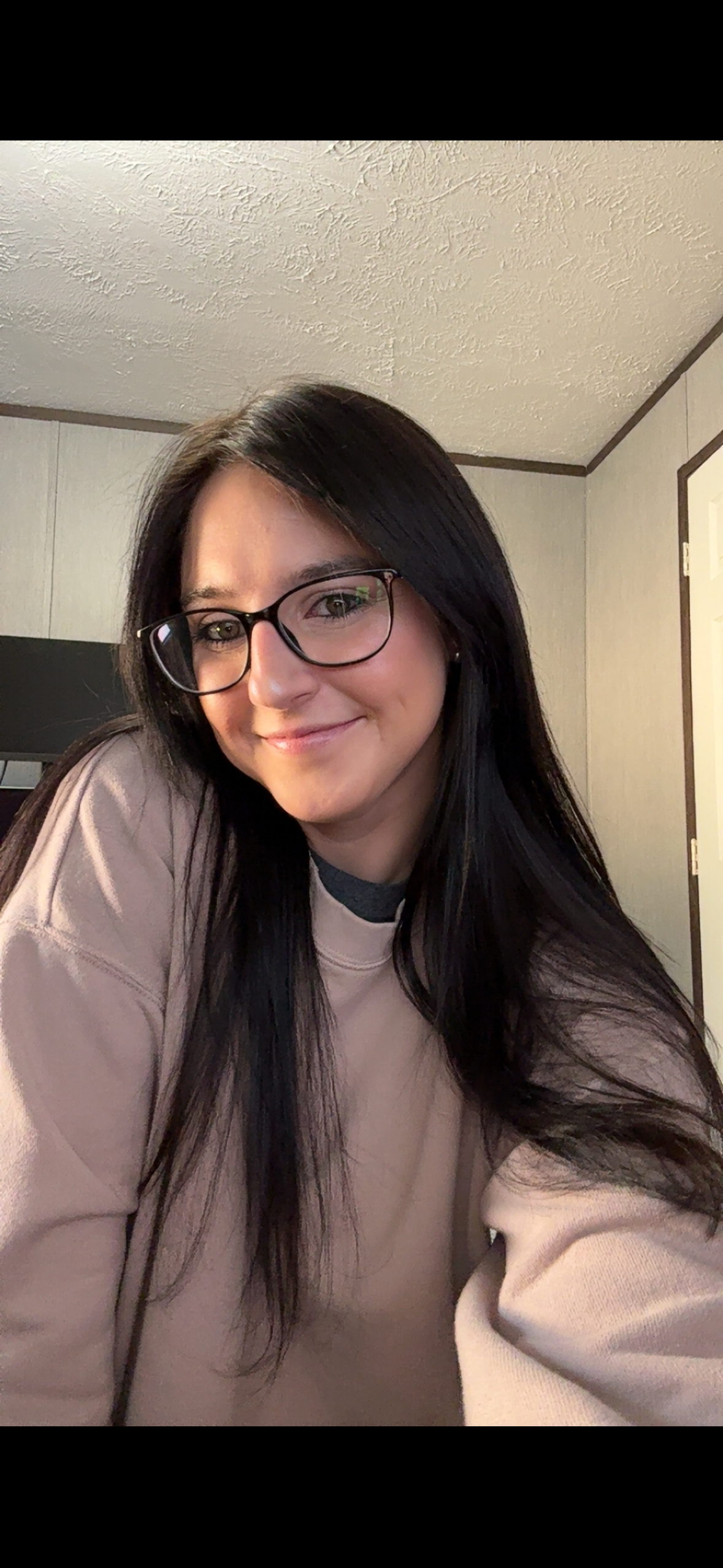 A woman with long dark hair and glasses smiling at the camera in a room with beige walls and a textured ceiling.