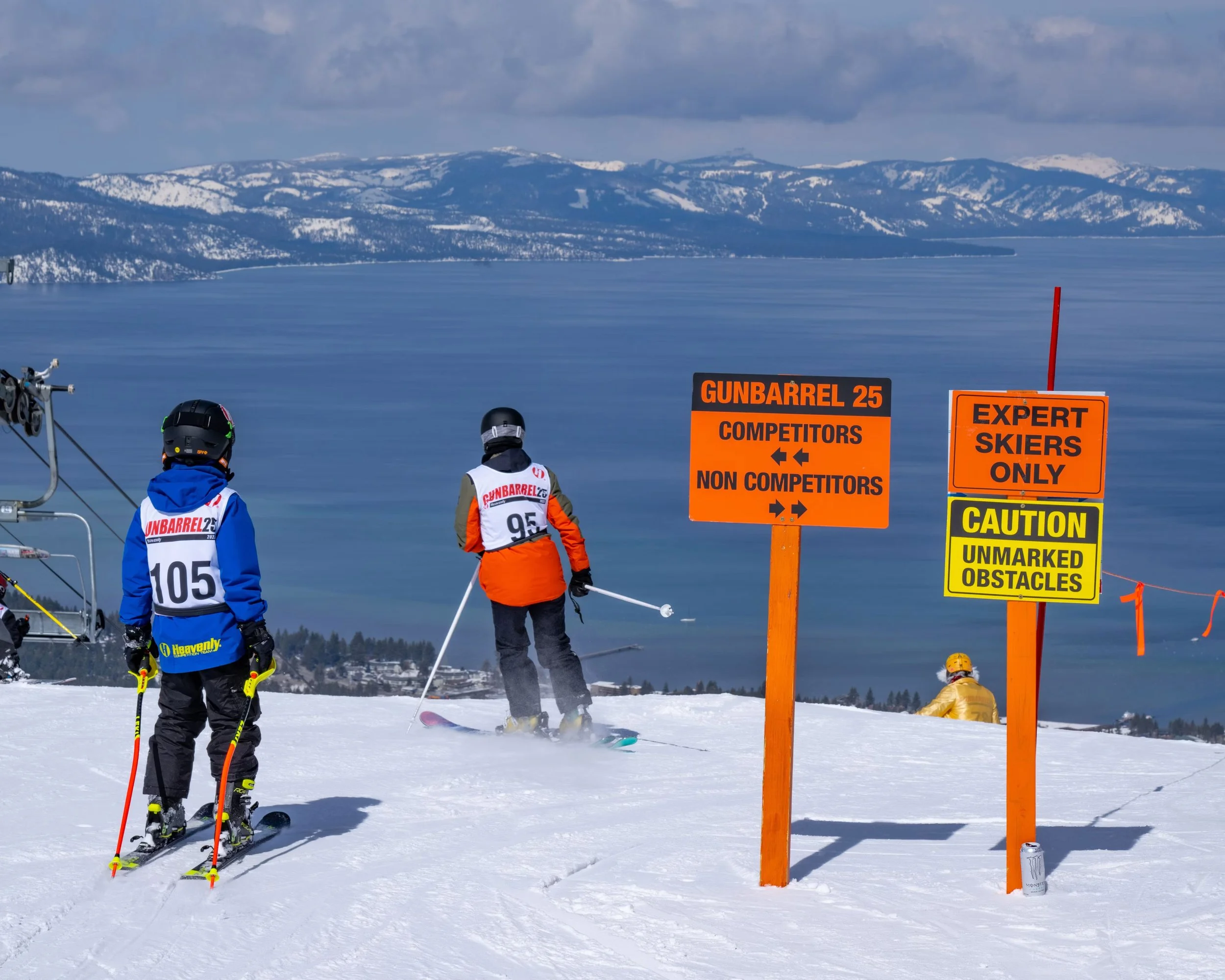 Skier in orange jacket and black pants preparing to ski down a snowy mountain, with two other skiers nearby, and three warning signs in orange and yellow about ski trail conditions and restrictions, over a landscape with snow-covered mountains and a body of water in the background.