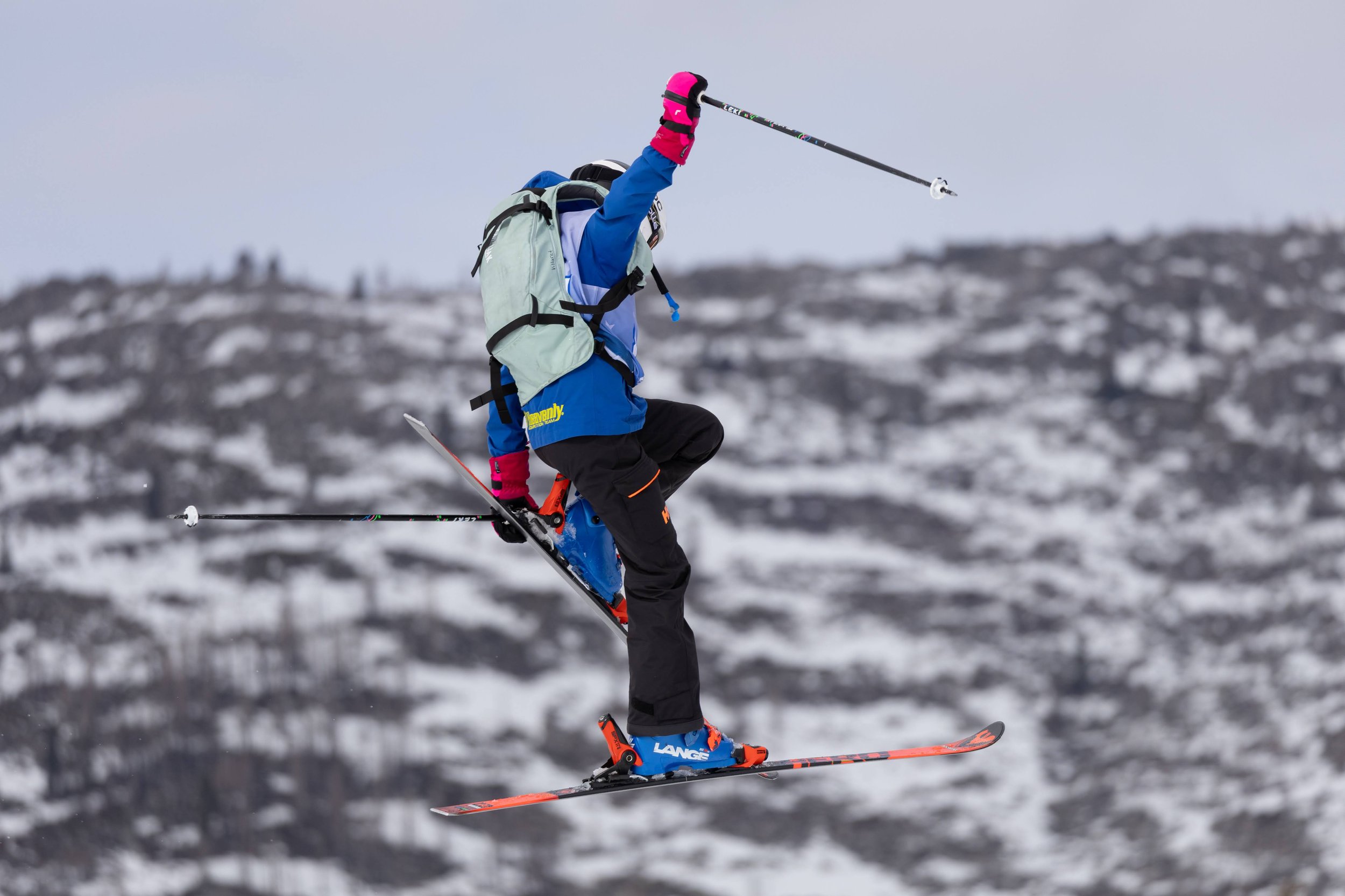 A person in colorful winter gear is mid-air on skis, with a snowy mountain landscape in the background.