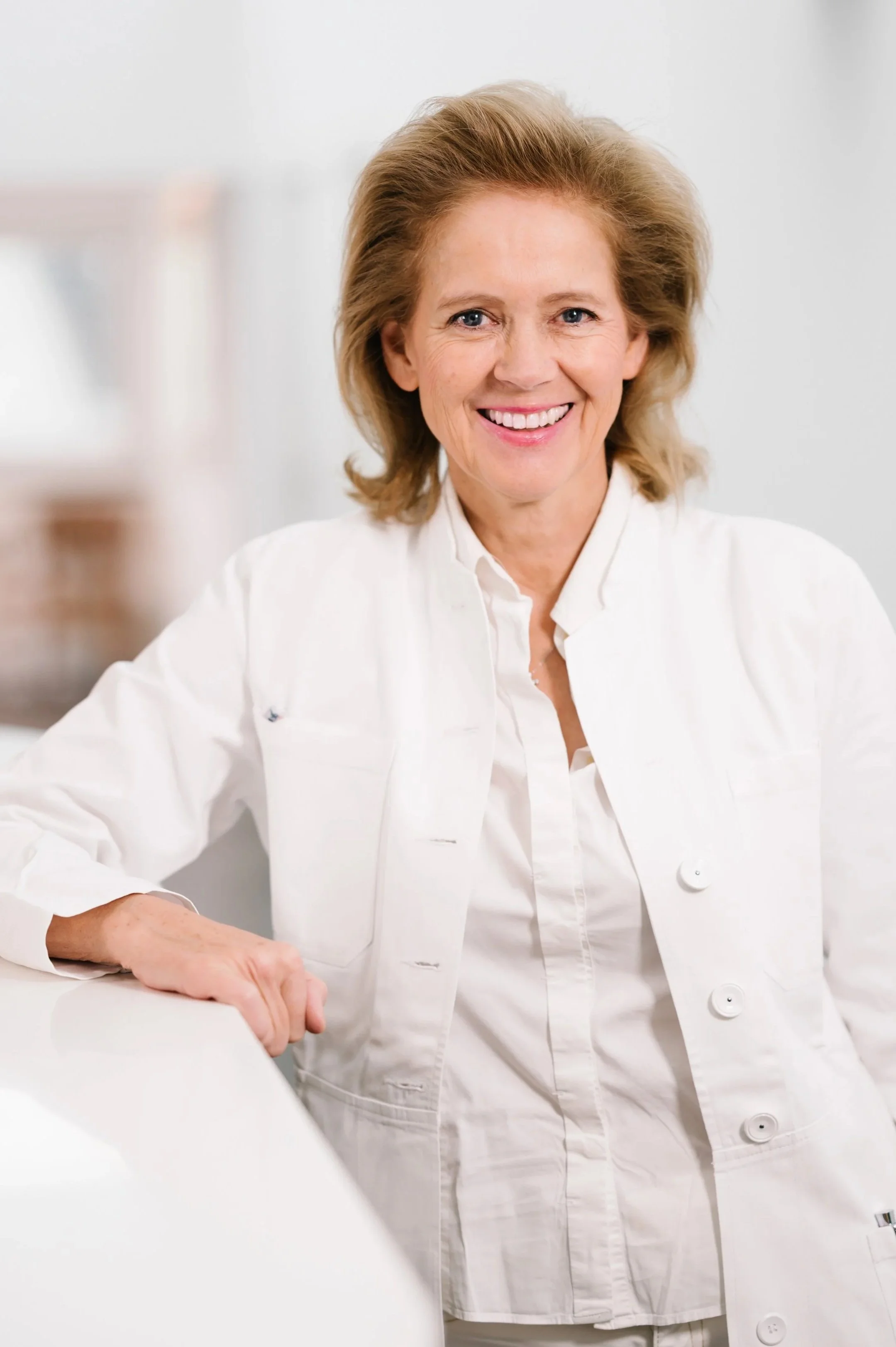 Eine lächelnde ältere Frau mit blonden Haaren trägt ein weißes Arzt- oder Pflegepersonal-Outfit und sitzt an einer weißen Oberfläche in einem hellen Raum.