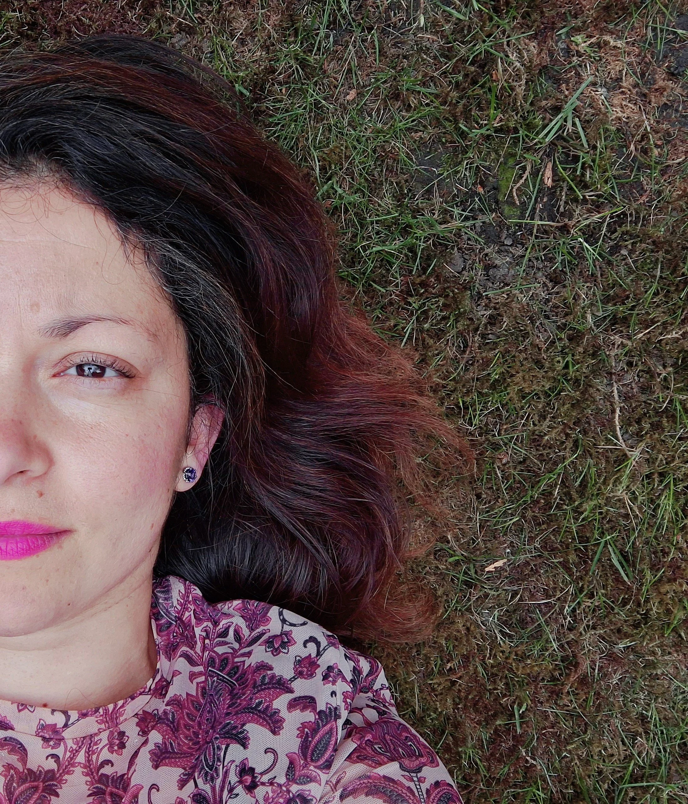 Partial portrait of a woman lying on the grass, visible from the shoulders up, with dark brown and reddish hair and wearing a paisley patterned blouse.