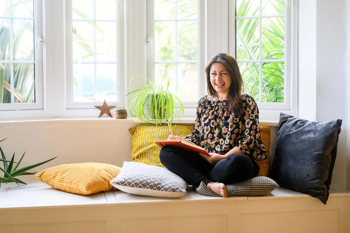 A woman sits cross-legged on a window seat, smiling and writing in a notebook, with large green plants and decorative pillows around her.