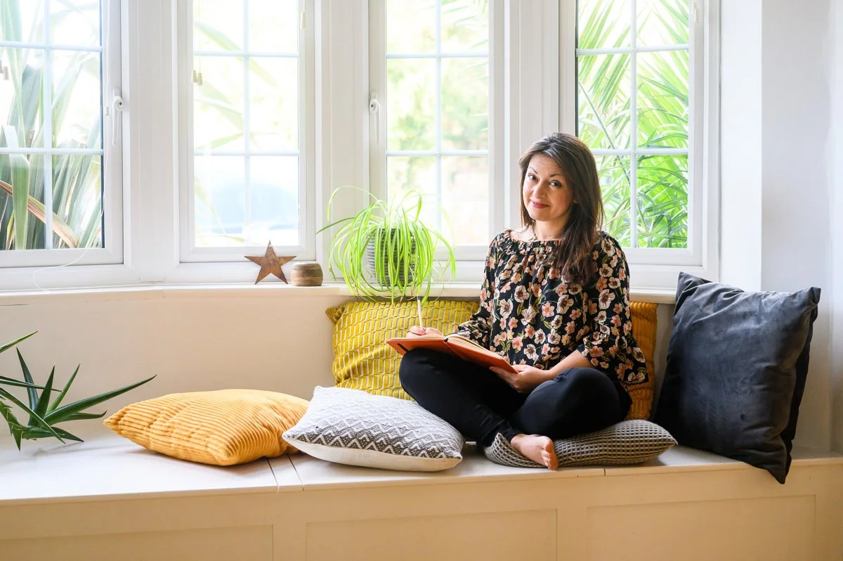 A woman sitting on a window seat with cushions, reading a book, with plants and outdoor greenery visible through the windows behind her.