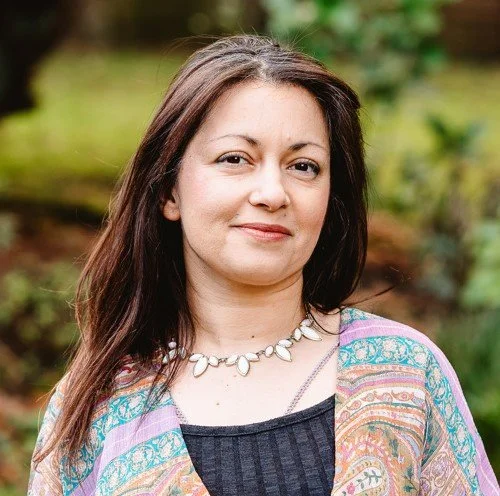 A woman with long brown hair outdoors wearing a colorful patterned shawl and a shell necklace.