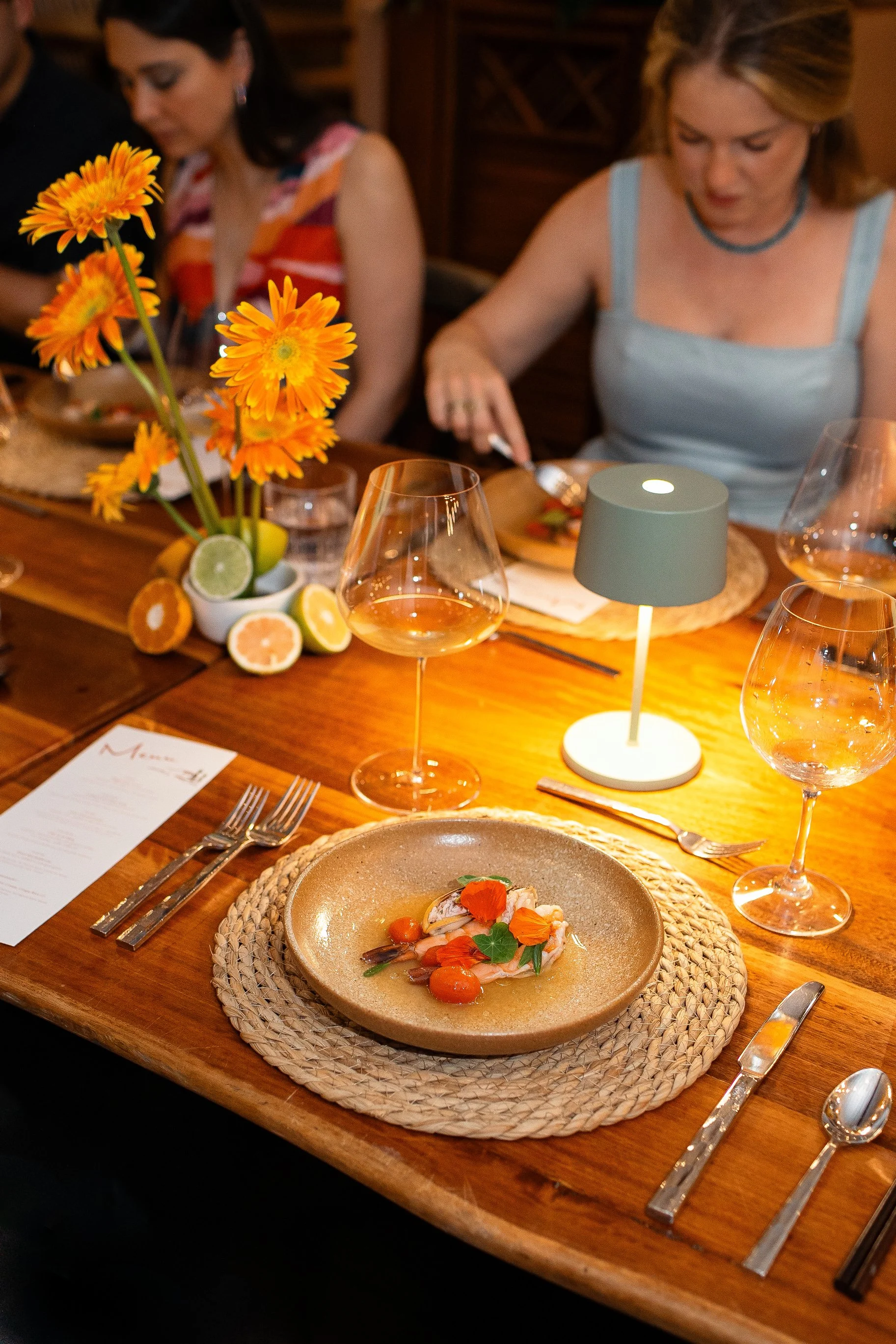 Elegant dinner table setting with a beige plate containing a gourmet dish garnished with orange and green edible flowers, a woven placemat, silverware, wine glasses, a vase with bright orange flowers and lemon and lime halves, and a small gray table lamp in a warmly lit restaurant.