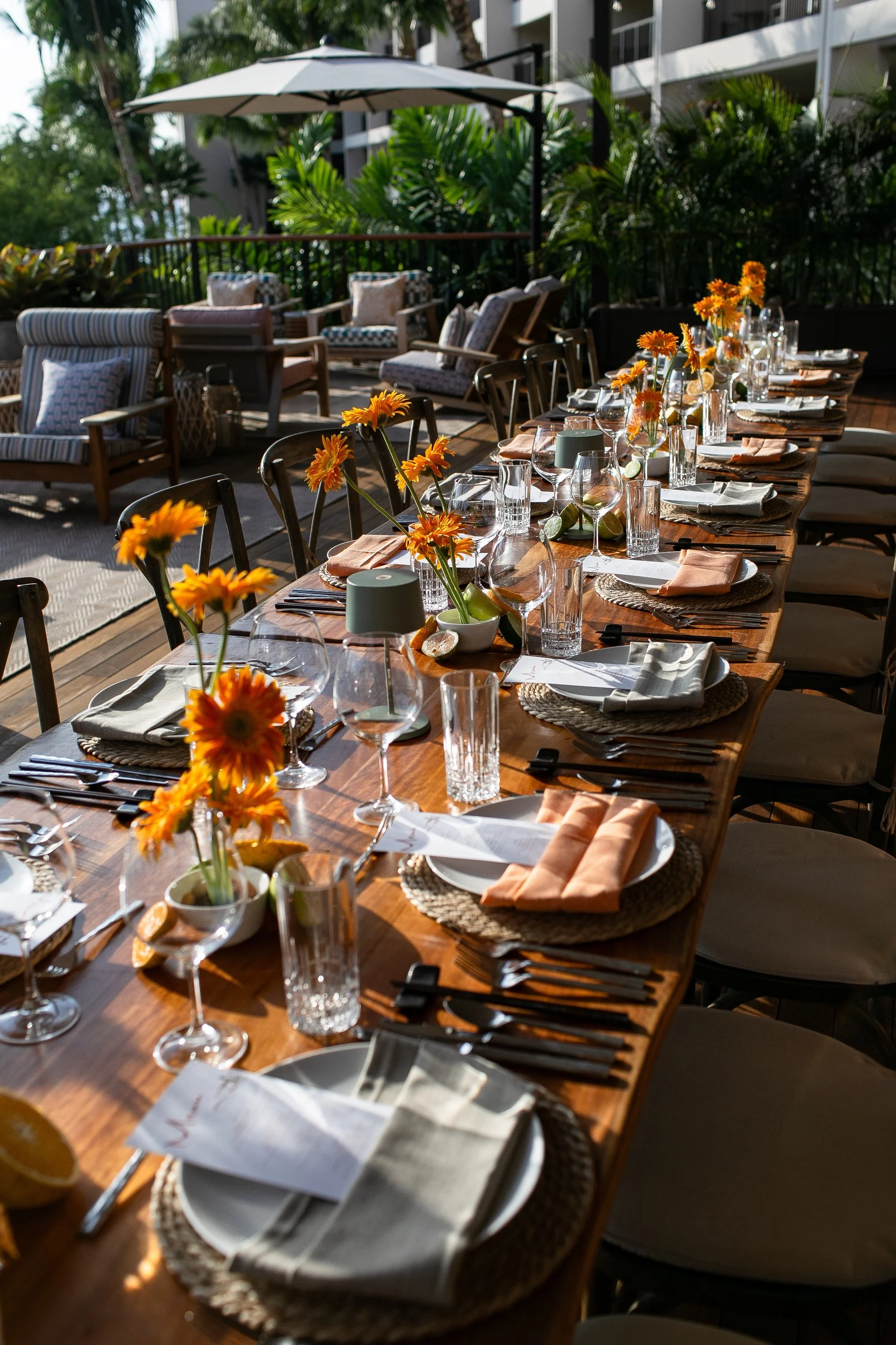 Outdoor dining table set with white plates, glassware, silverware, cloth napkins, and small floral arrangements of orange flowers, with a background of lounge chairs, cushions, a large umbrella, and lush greenery.