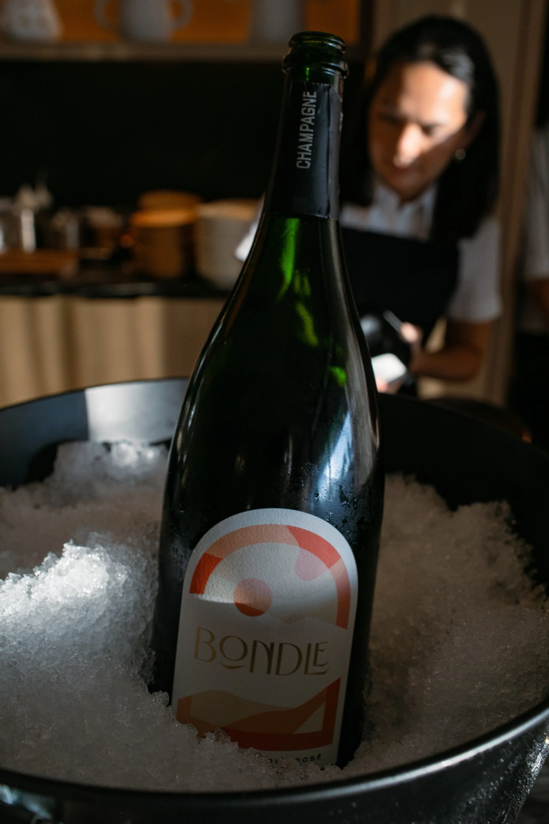 A bottle of BONDLE Champagne chilling in ice in a metal bucket with Chef Duyen Ha in the background.
