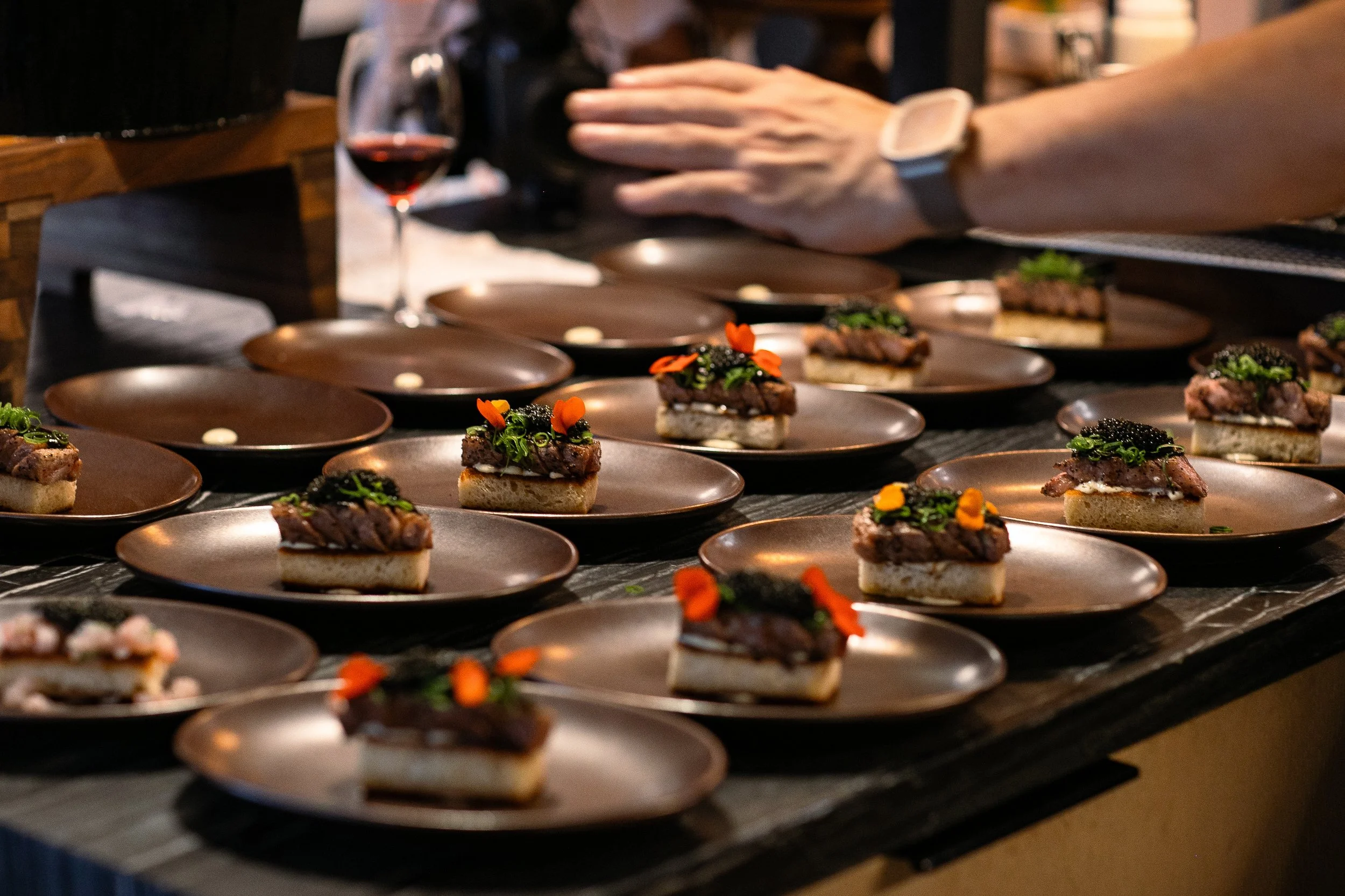 Multiple plated appetizers on a dark counter, with one hand reaching for a plate. The appetizers include small pieces of toasted bread topped with meat, green onions, black caviar, and edible orange flowers. A glass of red wine is visible in the background.