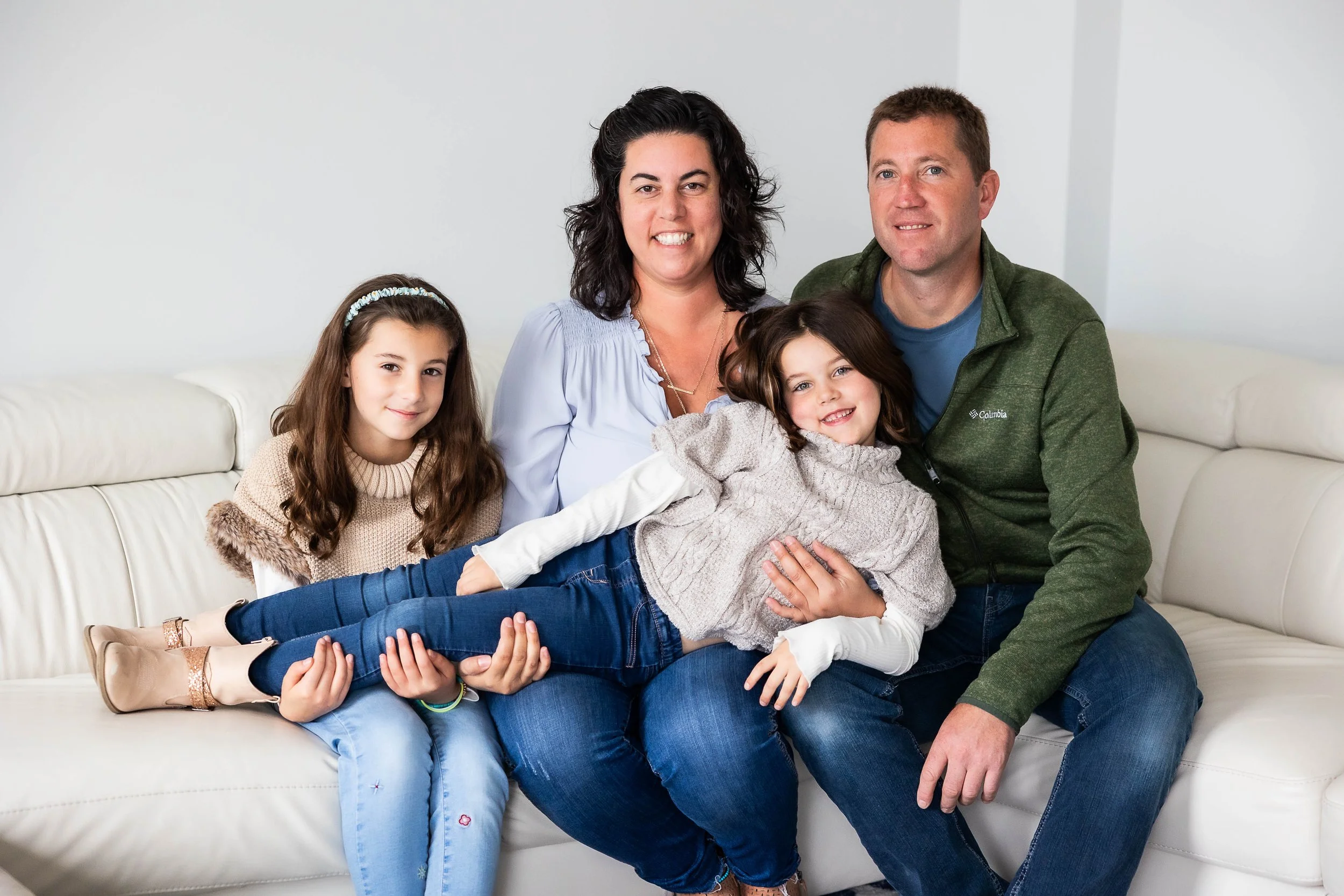 Family of five sitting on a couch, with two young girls and two adults, smiling and enjoying time together.