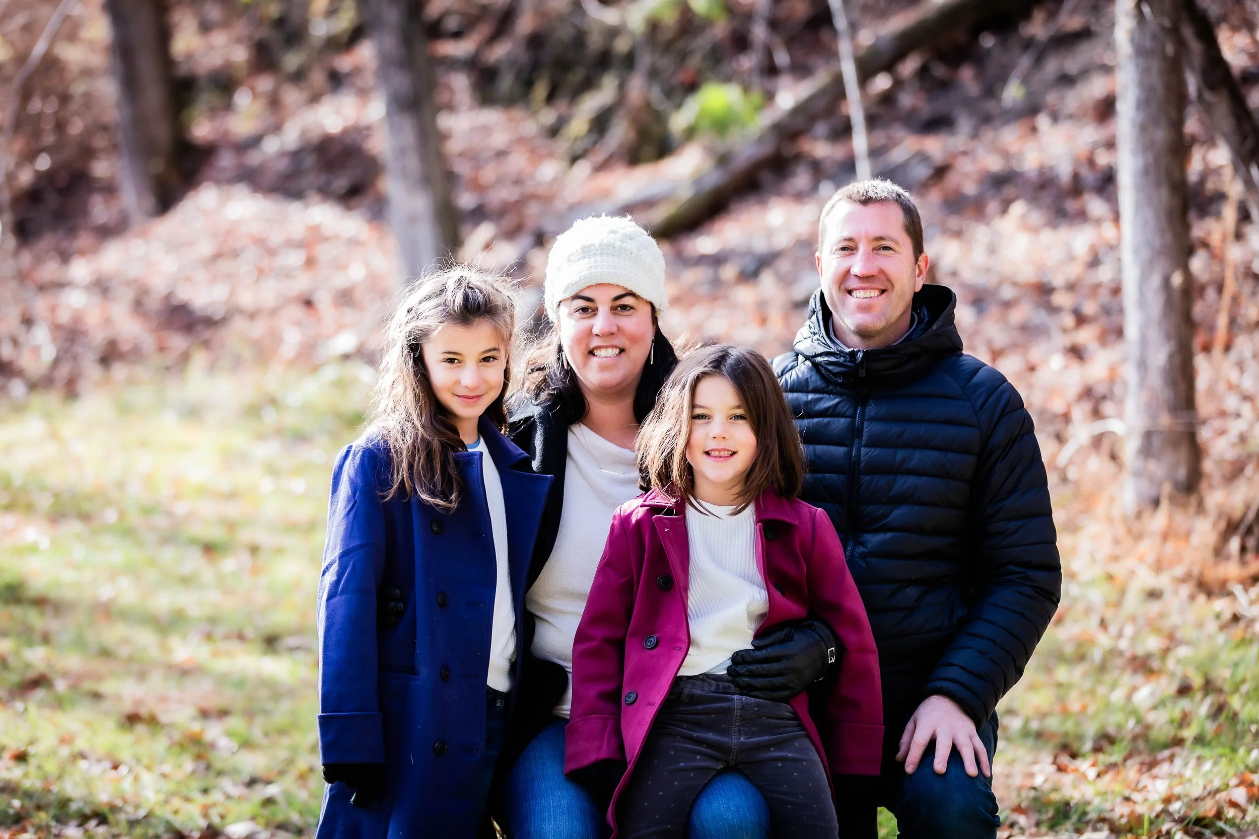 Family of four outdoors in autumn, smiling at camera, with trees and fallen leaves in background.