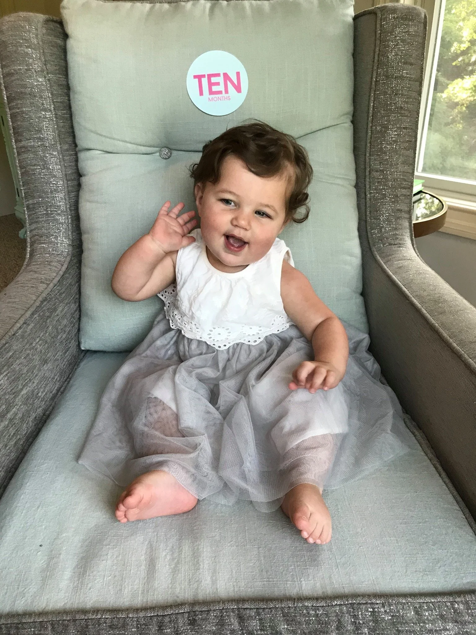A young girl sitting on a cushion on a chair, smiling and waving. A sticker with the text 'TEN MONTHS' is on the cushion behind her.