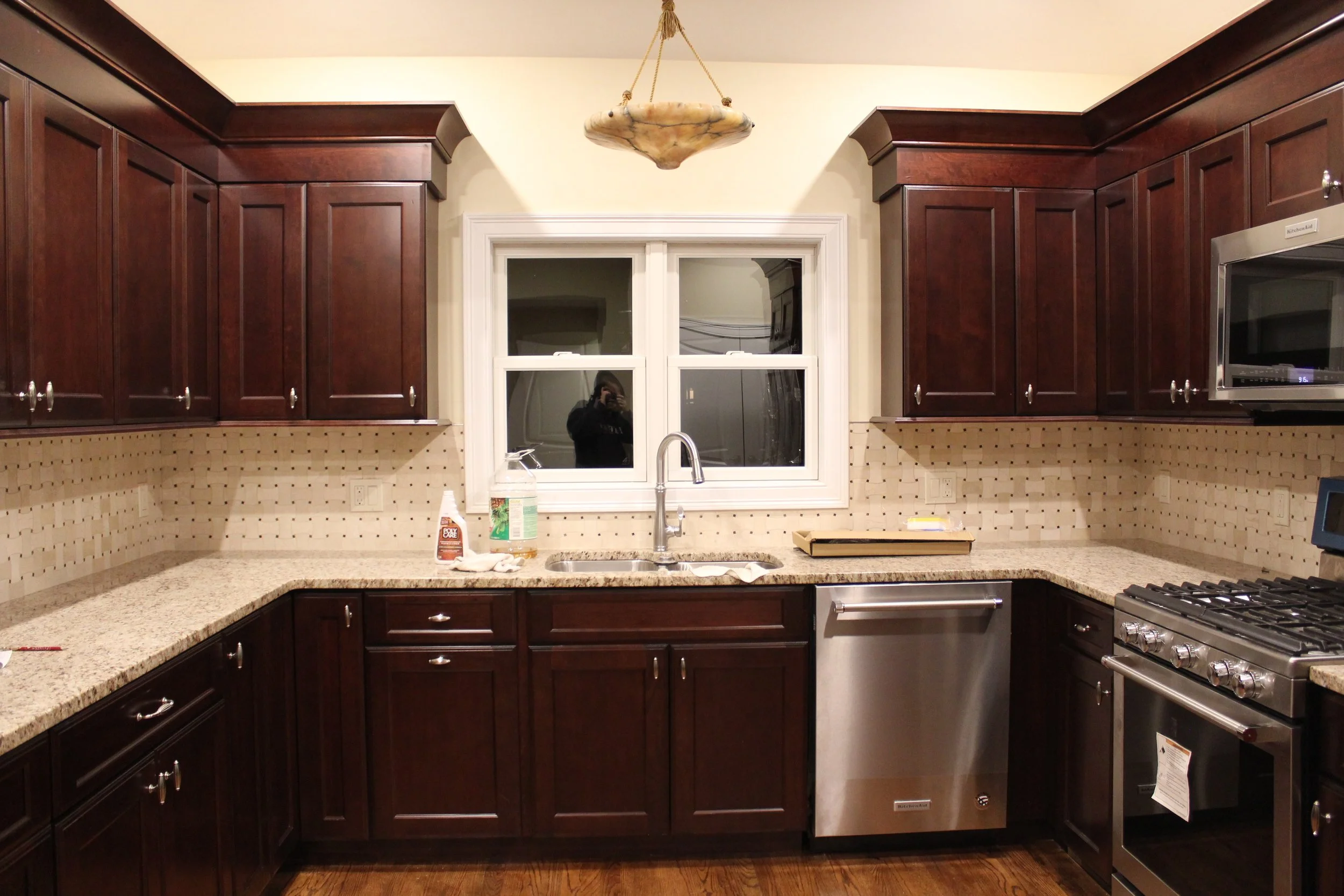 Kitchen with dark wood cabinets, beige speckled granite countertops, stainless steel dishwasher and stove, white window above the sink, and beige backsplash tiles with small black accents.