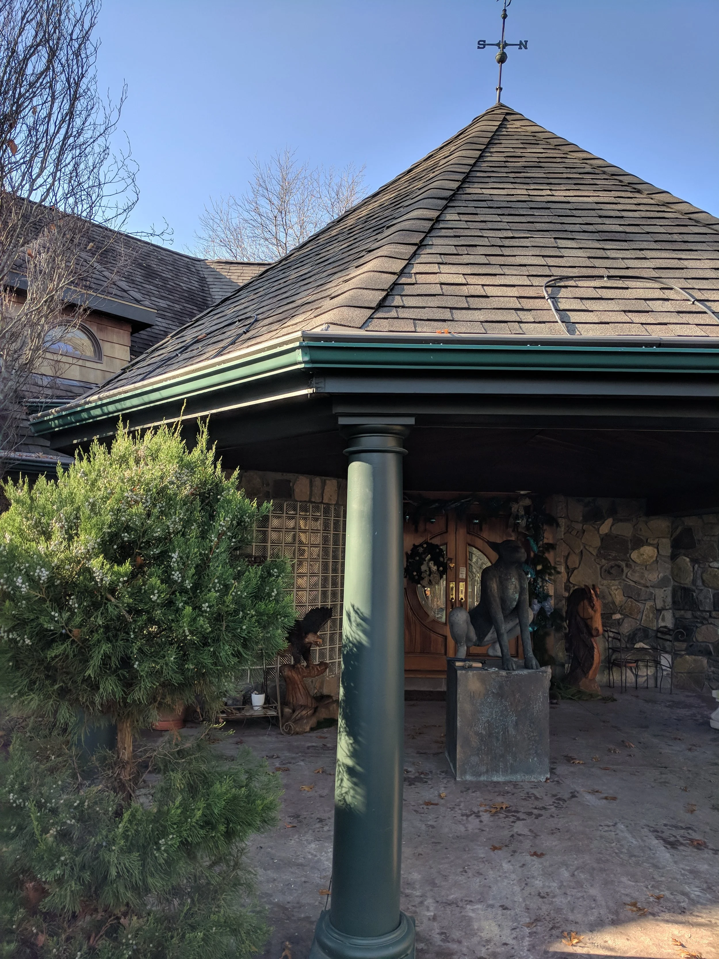 Front porch of a house with a sloped roof, stone wall, and column, decorated with sculptures, plants, and holiday wreaths.