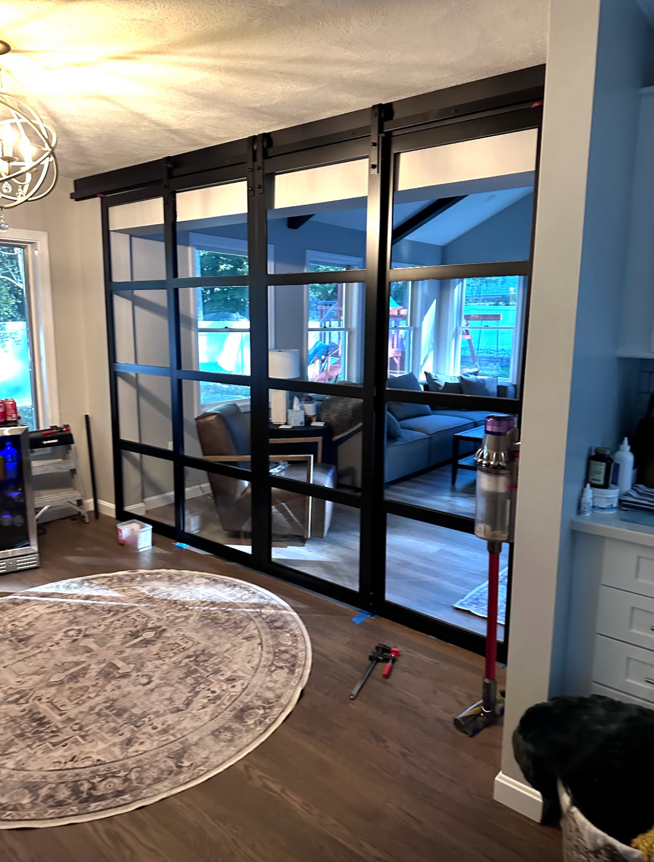 Living room with a glass panel room divider, furniture, window view of a backyard playground, and a vacuum cleaner near the divider.