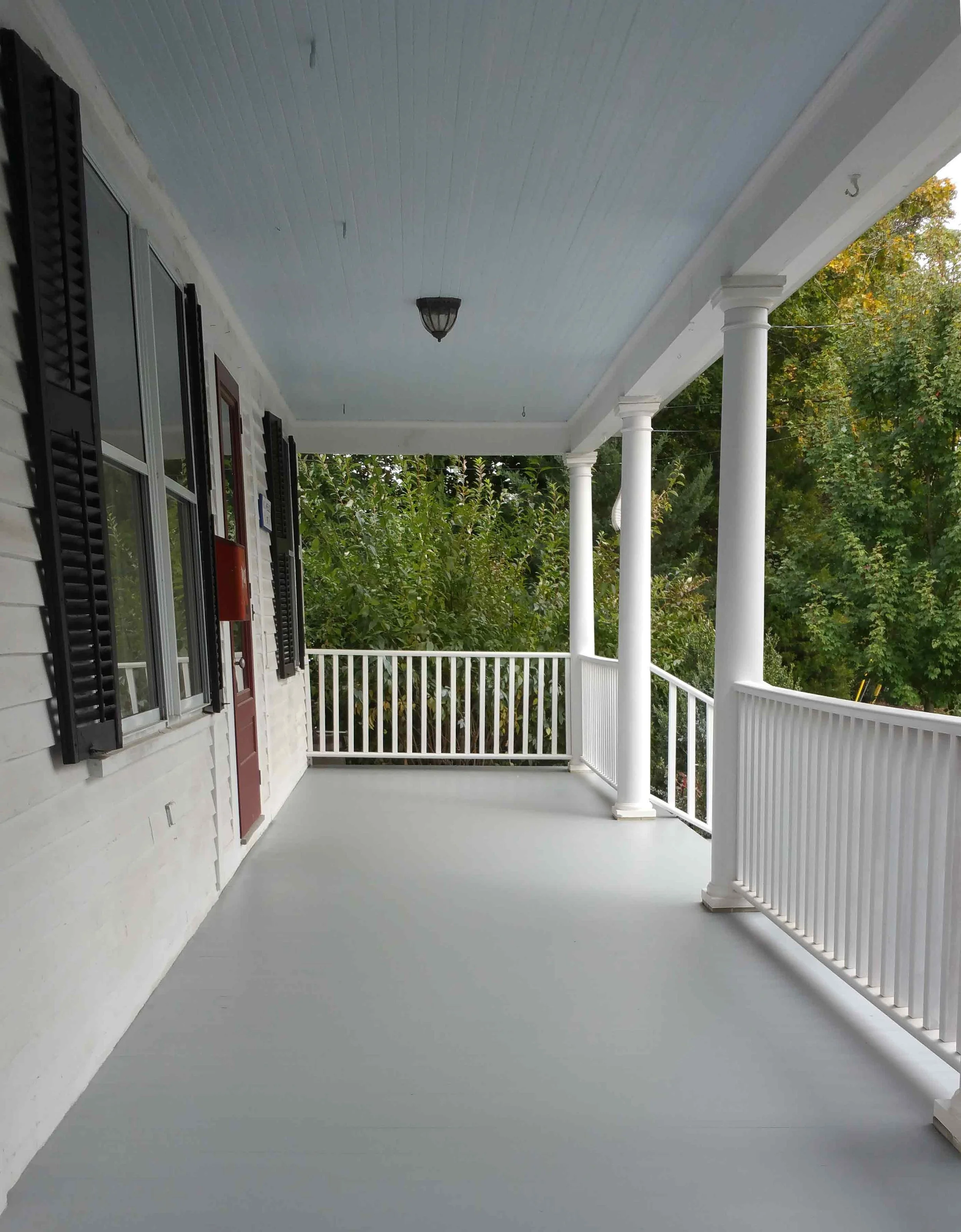 Empty porch with white railing, columns, and light blue ceiling, attached to a white brick house with black window shutters and a red door, surrounded by green trees.