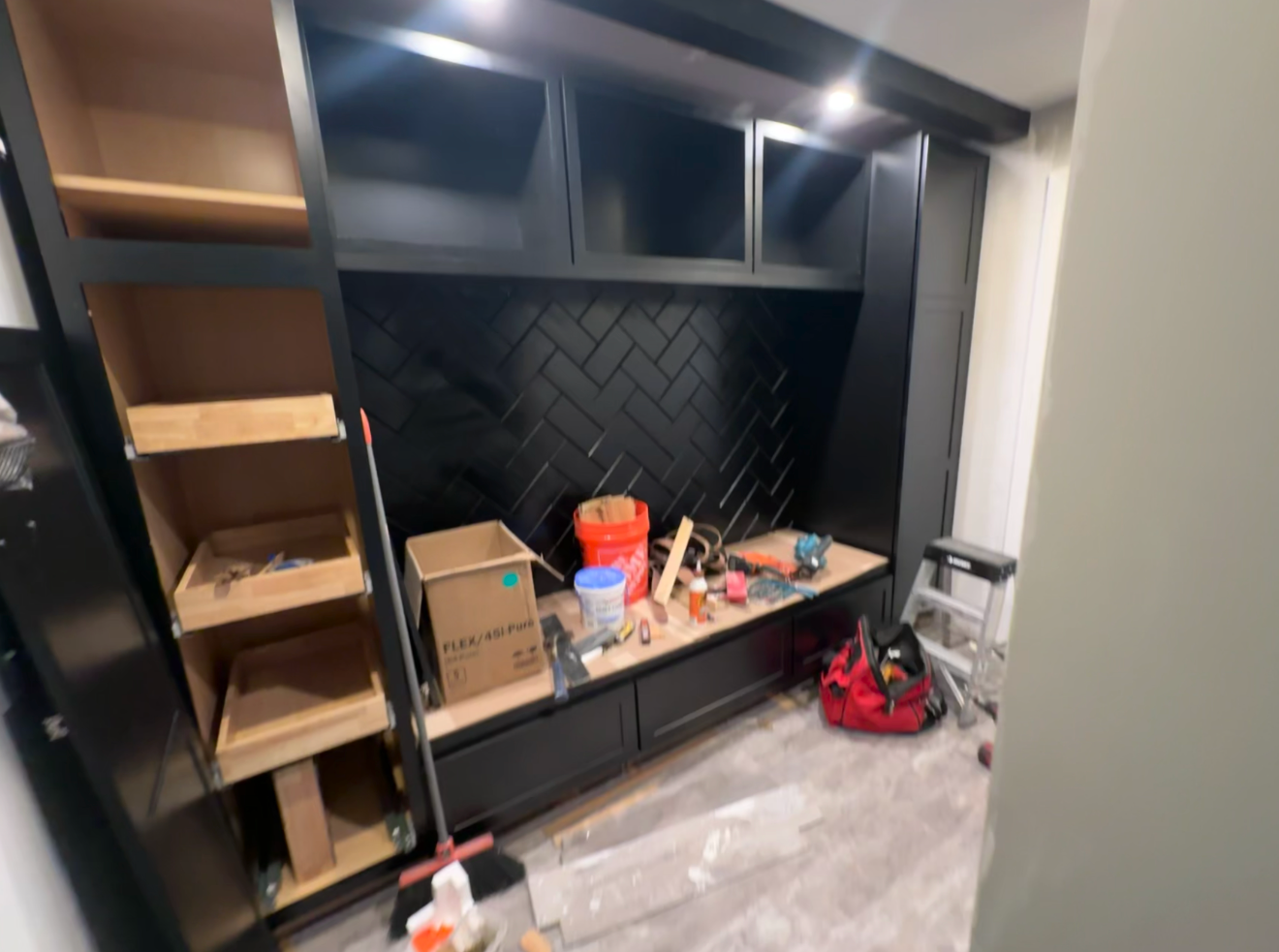 Kitchen cabinetry under construction with black cabinets, some open shelves, tile backsplash, tools, and construction materials on the countertop.