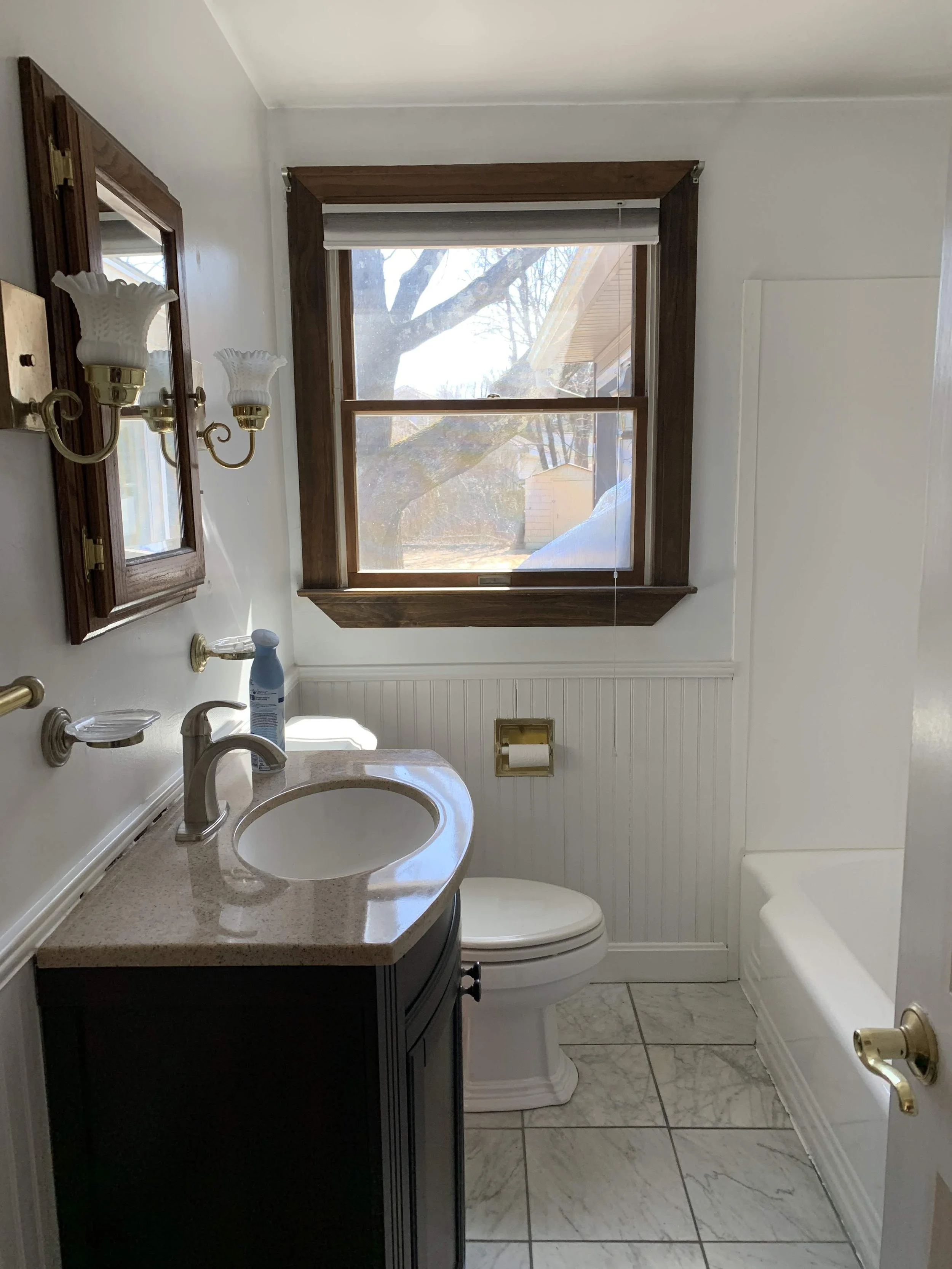 Small bathroom with white walls, a marble floor, a wooden framed window showing a tree outside, a white toilet, a dark vanity with a beige countertop, a mirror, and an old-fashioned brass toilet paper holder.