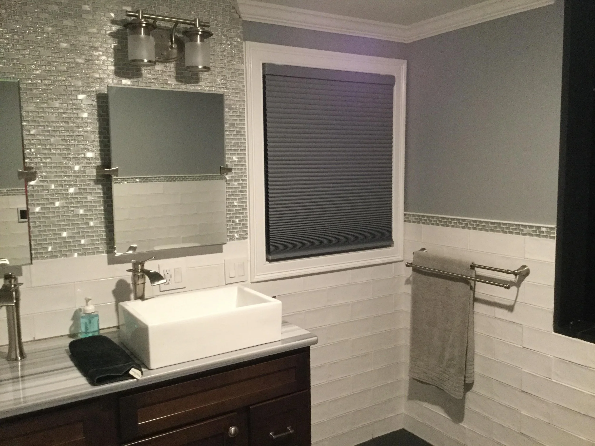 Modern bathroom with white brick walls, a dark wooden cabinet with a vessel sink, a mirror, a towel hanging on a towel bar, and a window with closed blinds.