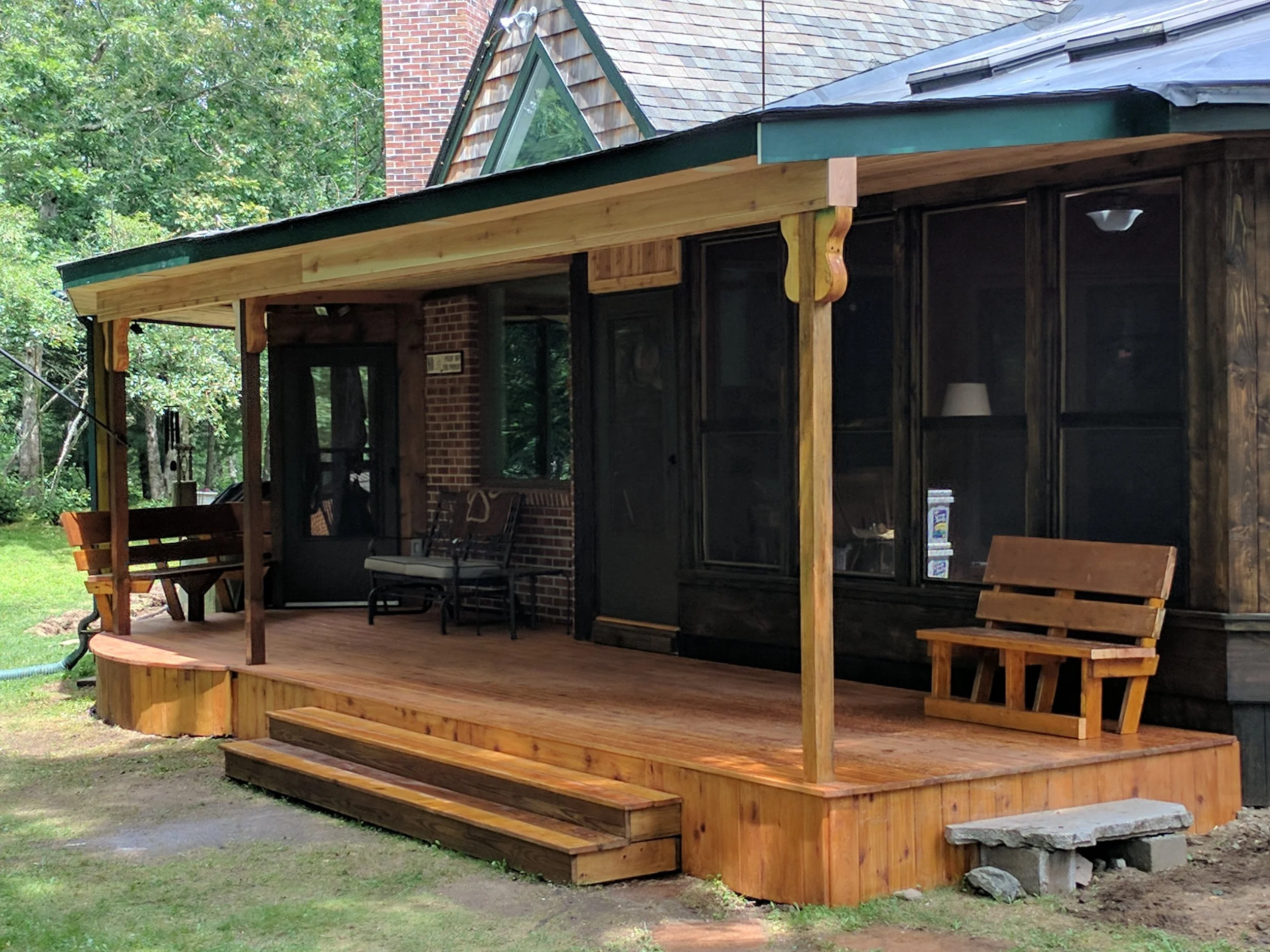 Wooden porch attached to a brick house with glass windows, outdoor chairs, a bench, and steps leading to the lawn, surrounded by green trees.